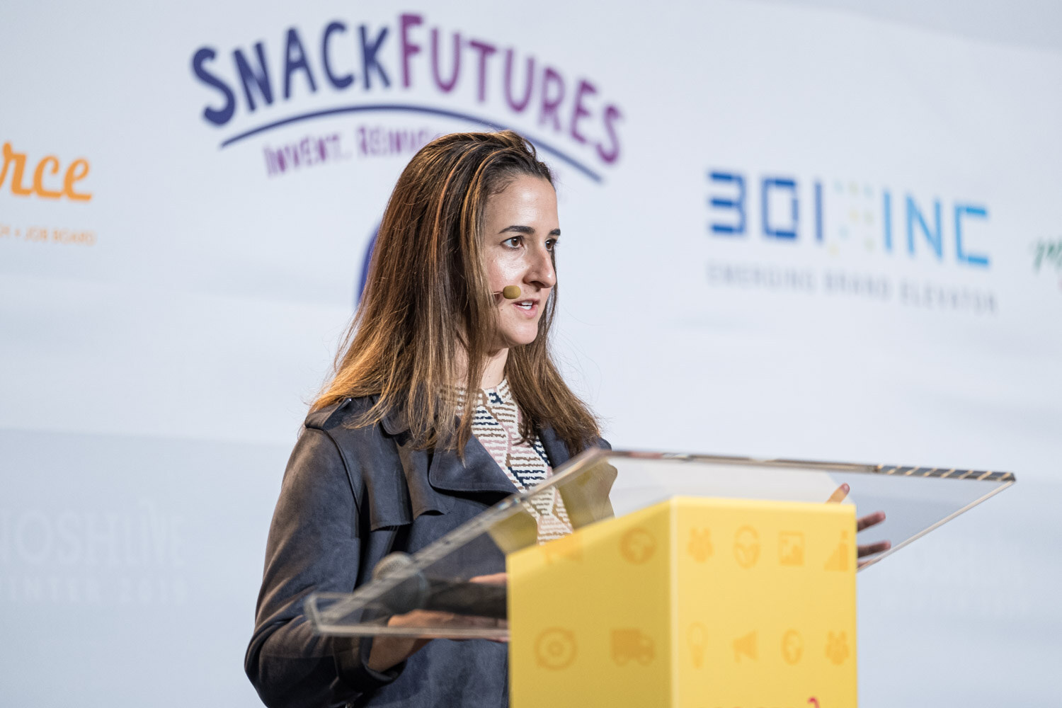 A woman with long hair at a yellow Nosh Live lectern in front of SnackFutures and BOI Inc banners
