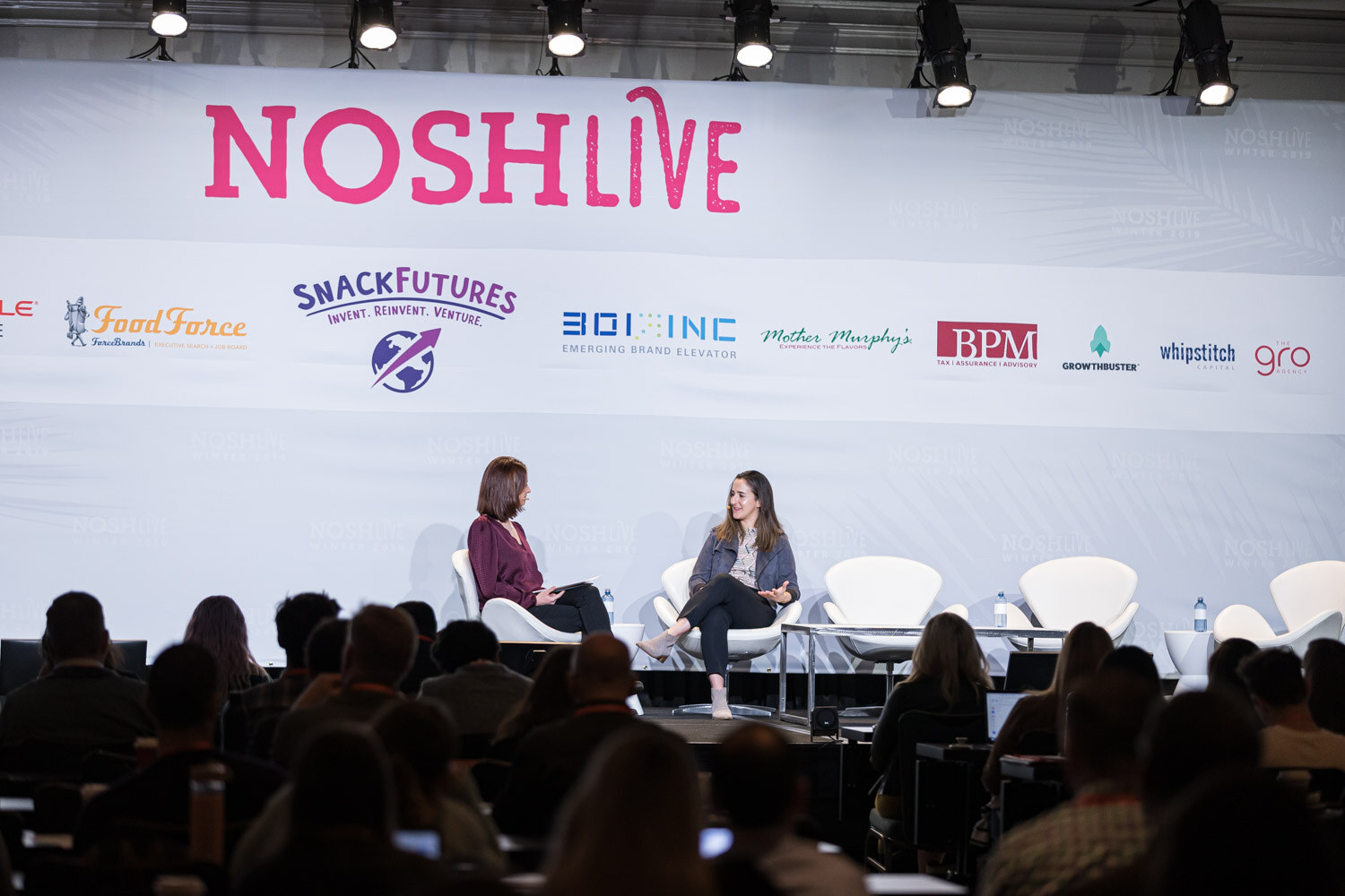 A moderator and a speaker seated on stage with the audience in silhouette in front