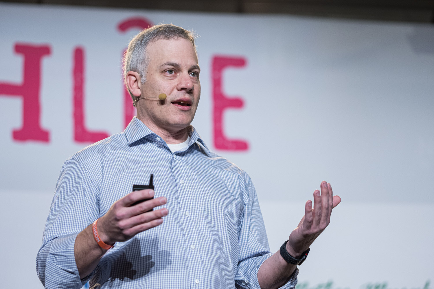A man in a light blue checked shirt with a headset microphone gesturing in front of the Nosh Live logo