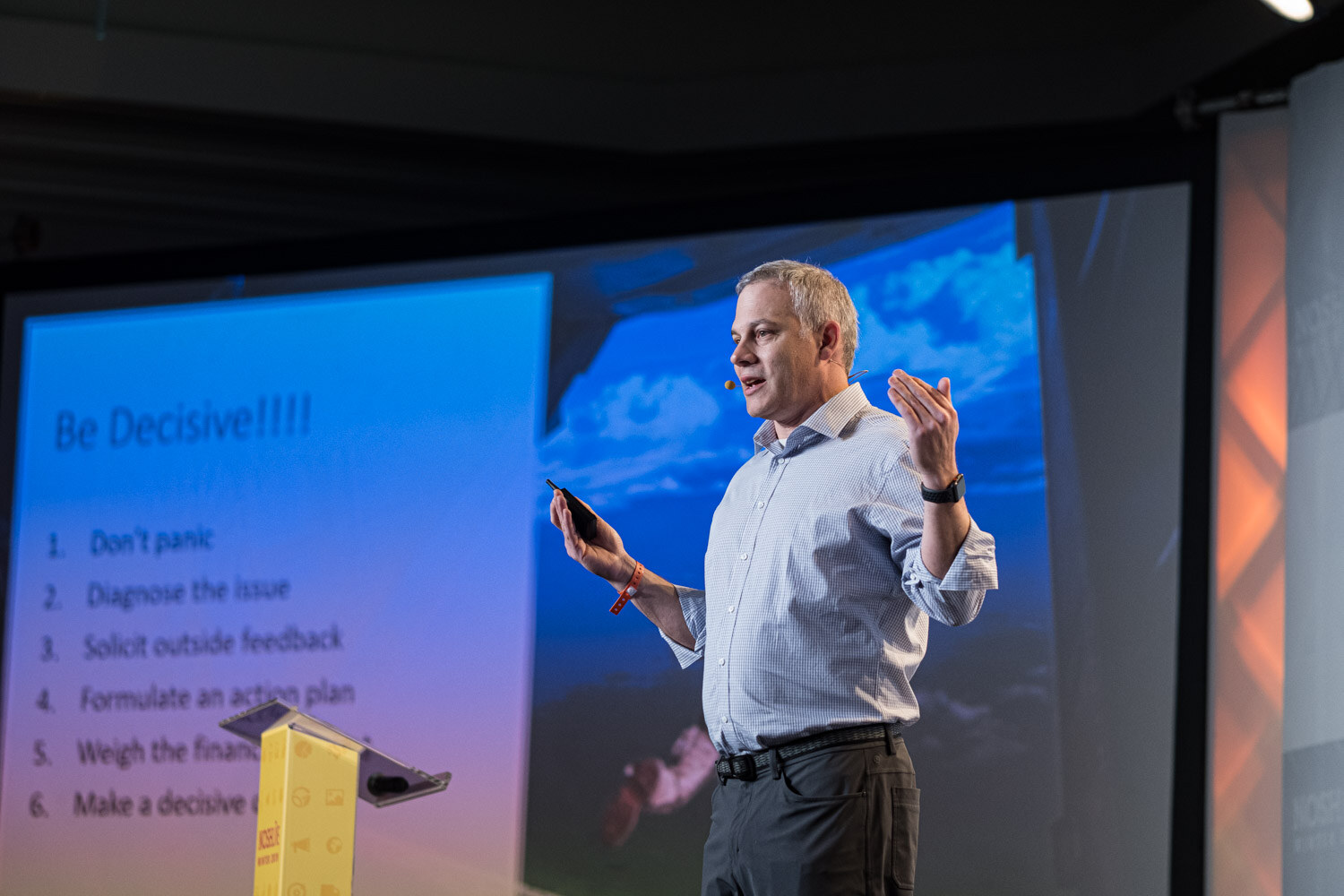 A man on stage beside a slide titled Be Decisive with a numbered list