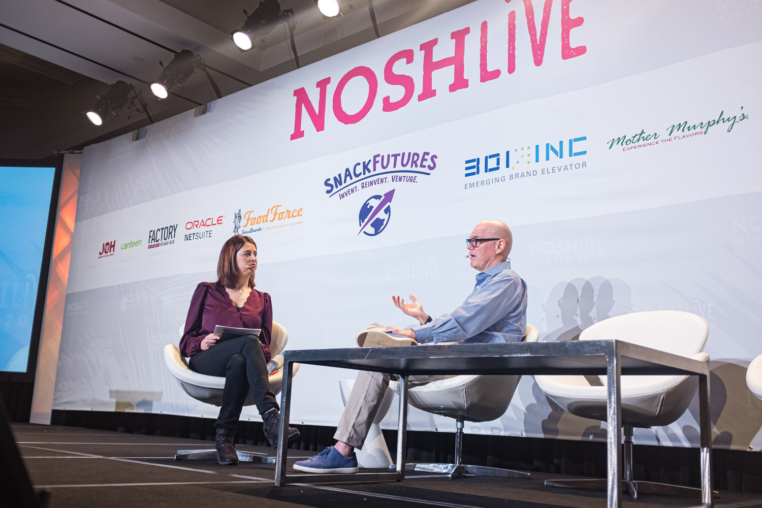Two people seated in chairs on stage under the Nosh Live banner and sponsor logos
