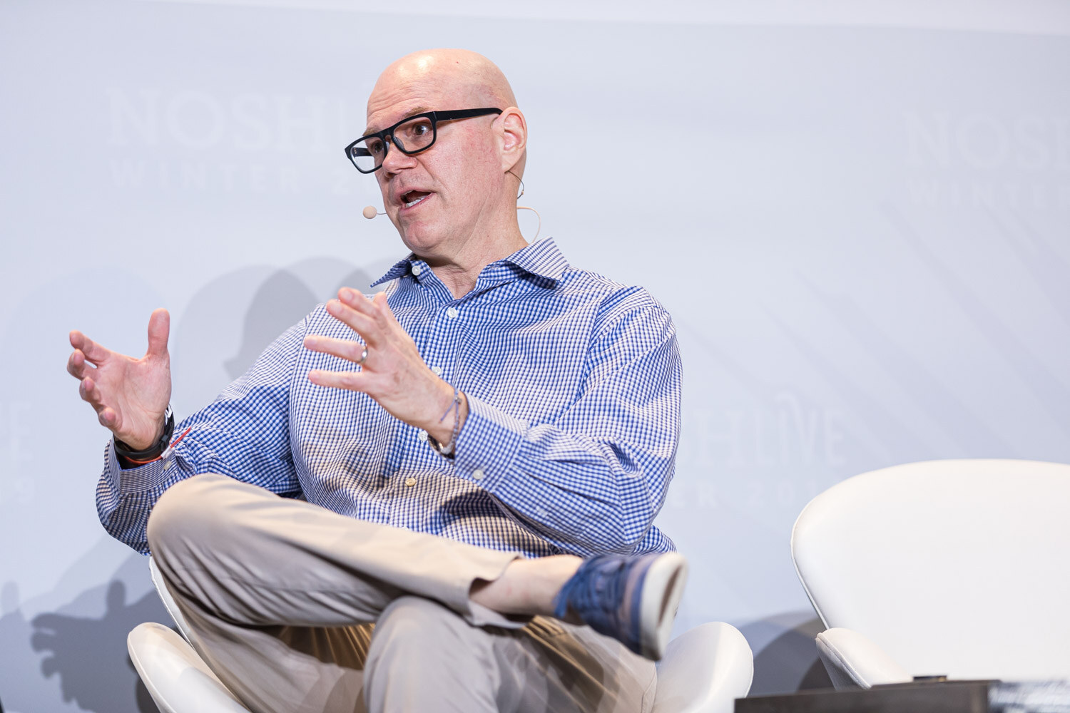 A man in glasses and a blue checked shirt gesturing with both hands while seated
