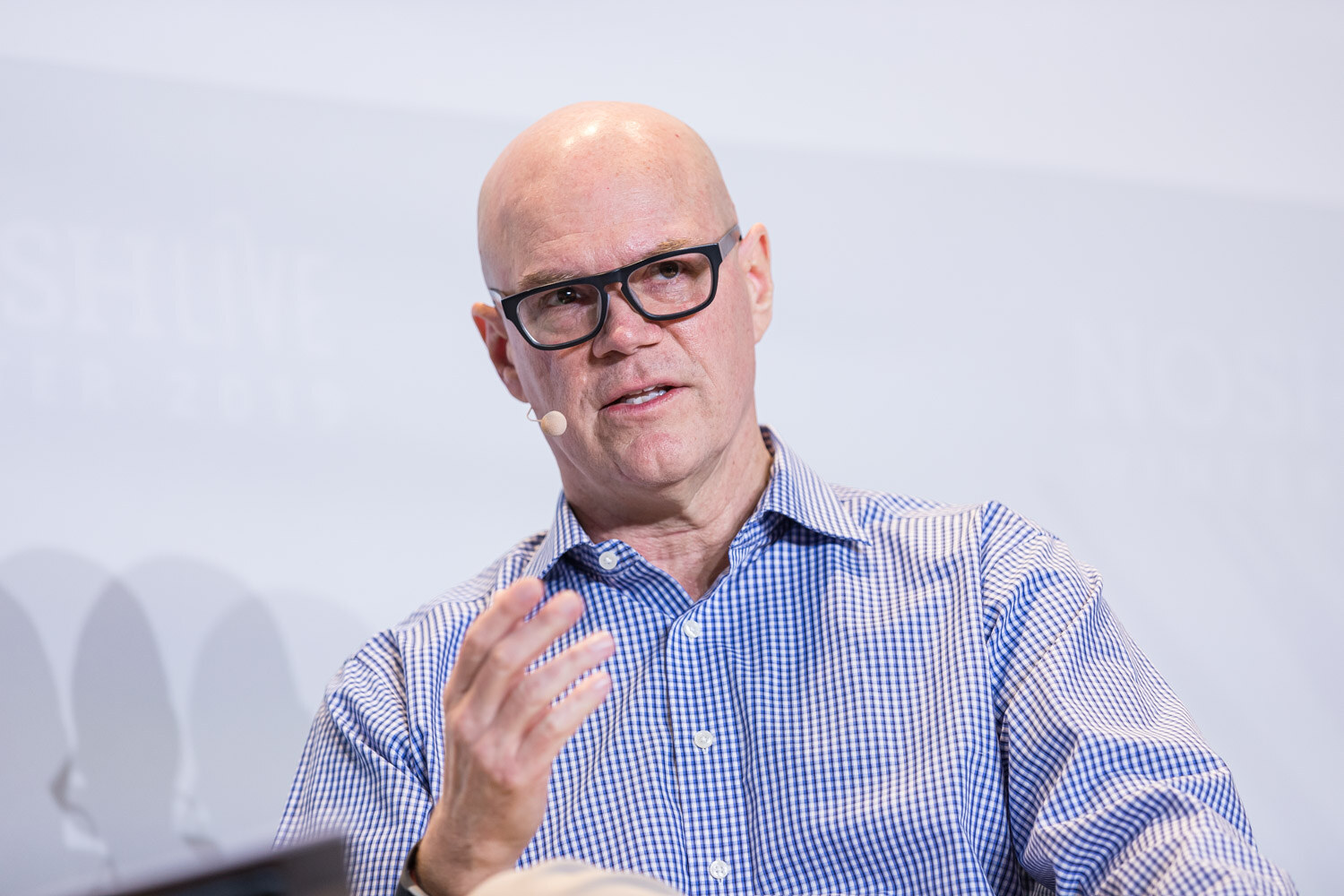 A bald man in black-framed glasses speaking with one hand raised