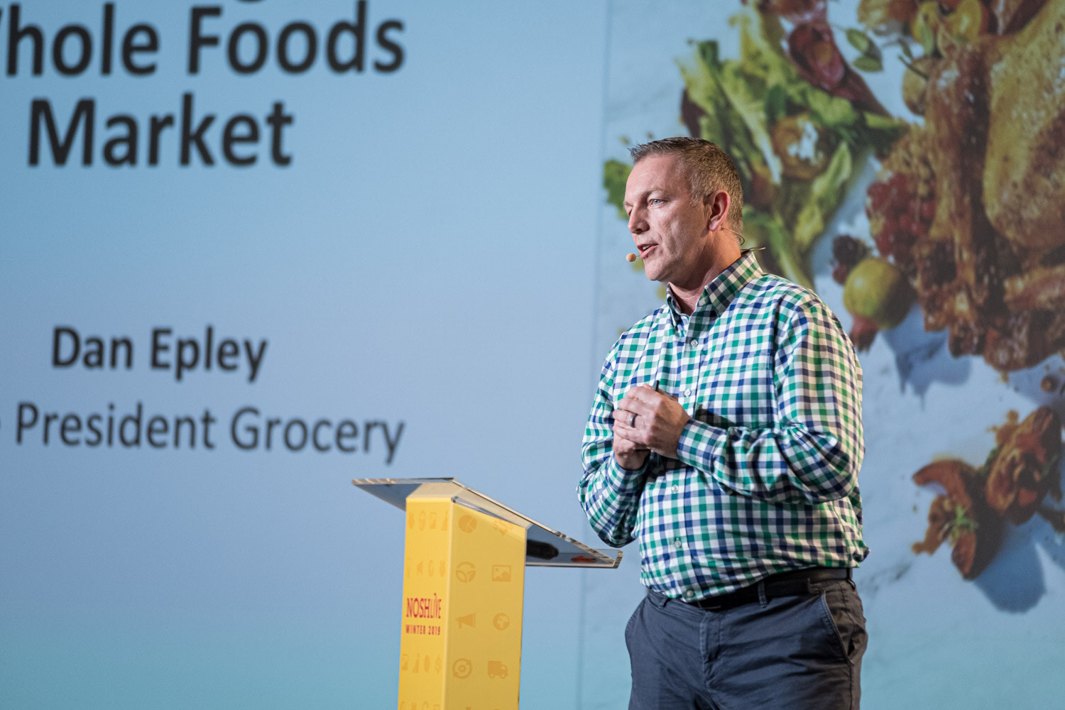 A man in a green checked shirt at a lectern in front of a slide naming Dan Epley, President Grocery, Whole Foods Market