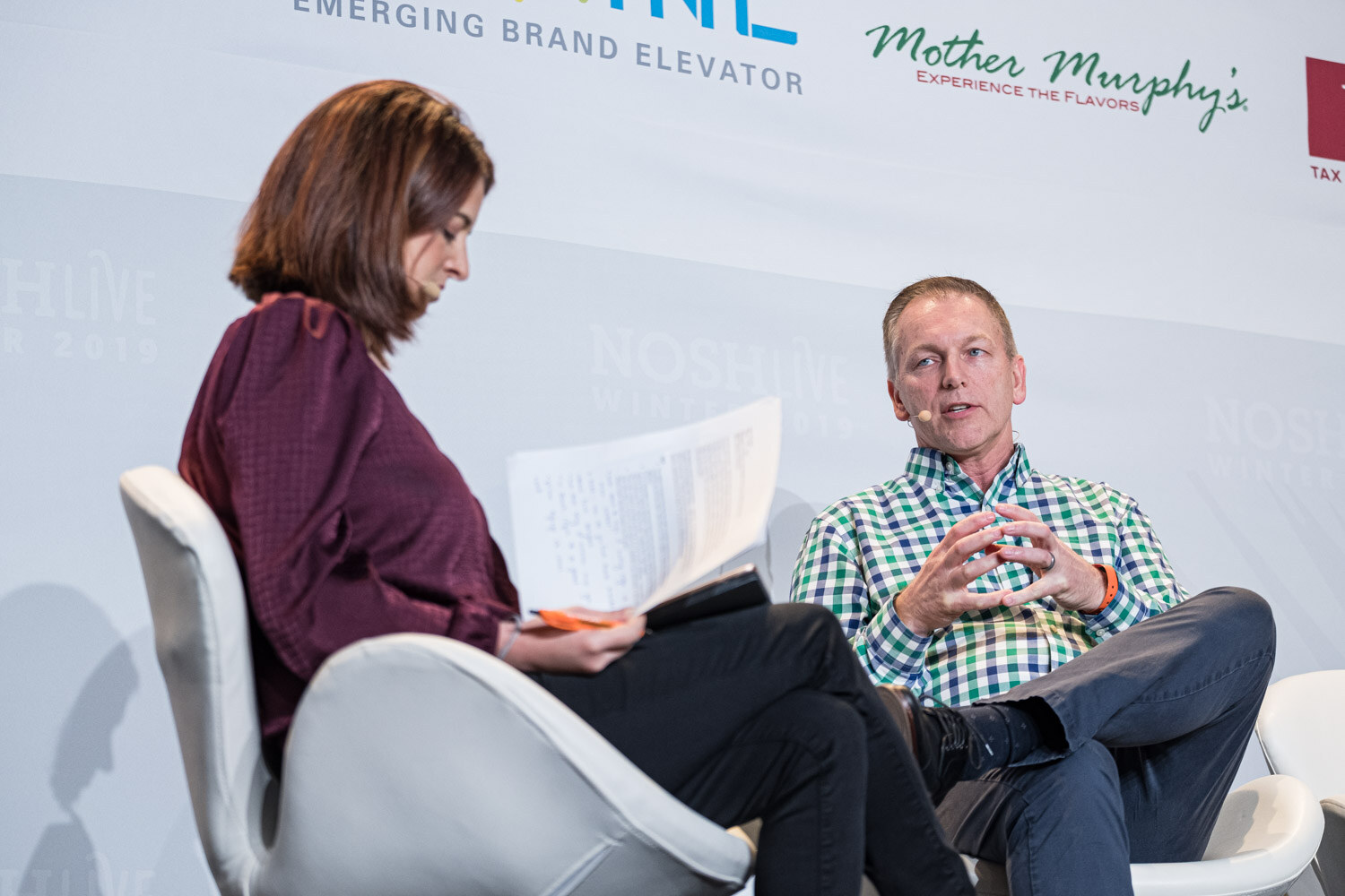 A moderator holding papers seated beside a man in a green checked shirt with hands clasped