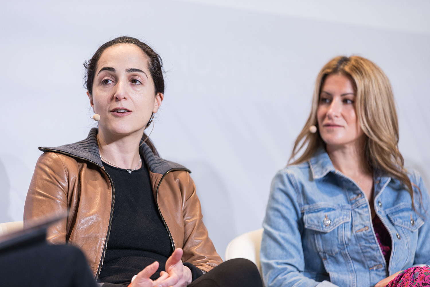 A woman in a brown leather jacket speaking while a woman in a denim jacket looks on