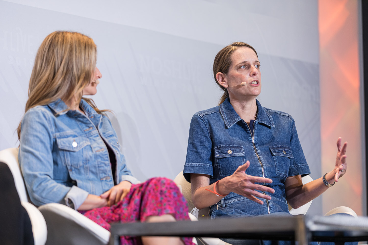 A woman in a denim jumpsuit gesturing as she speaks next to a woman in a denim jacket