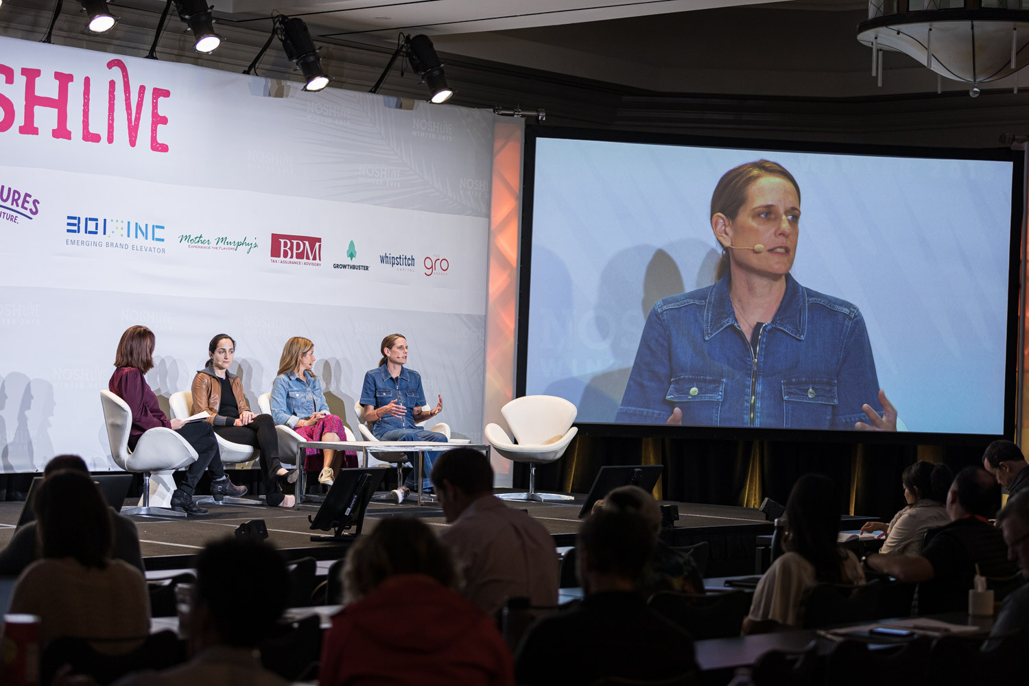 The four-person panel on stage while a large screen shows a close-up of one panelist