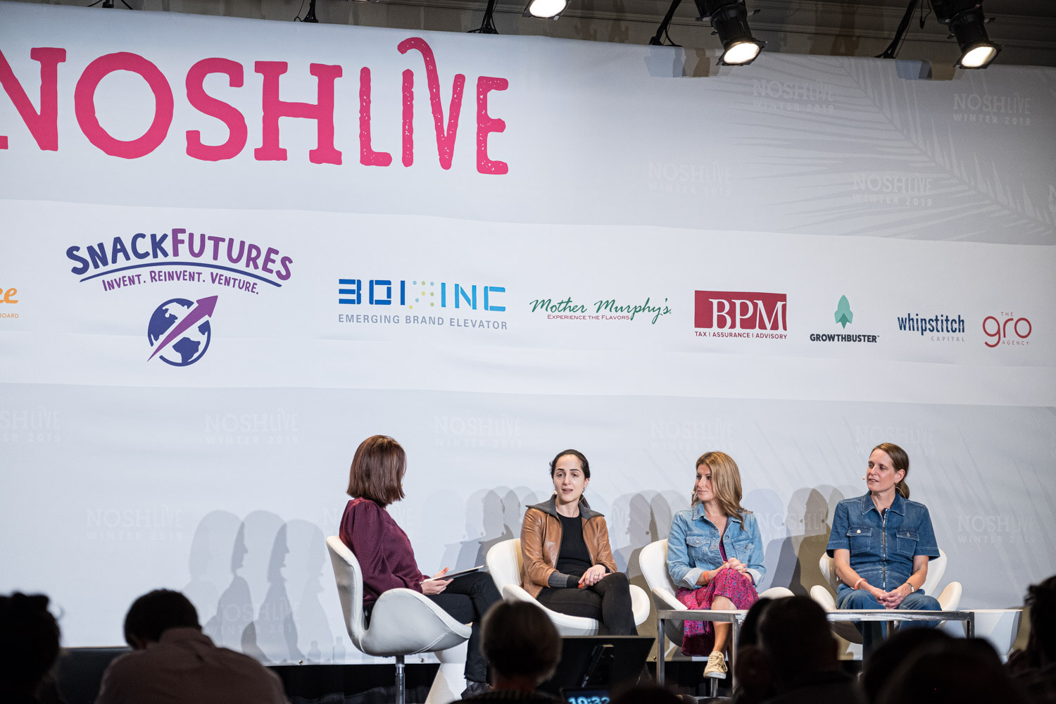 A moderator and three panelists seated in white chairs under the Nosh Live banner with the audience in silhouette