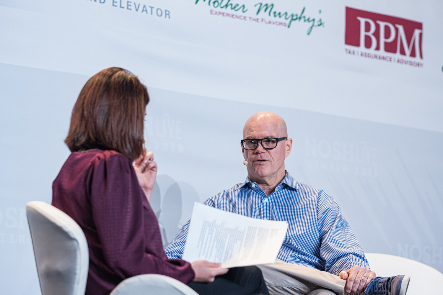 A moderator with a printout seated across from a bald man in glasses and a blue checked shirt