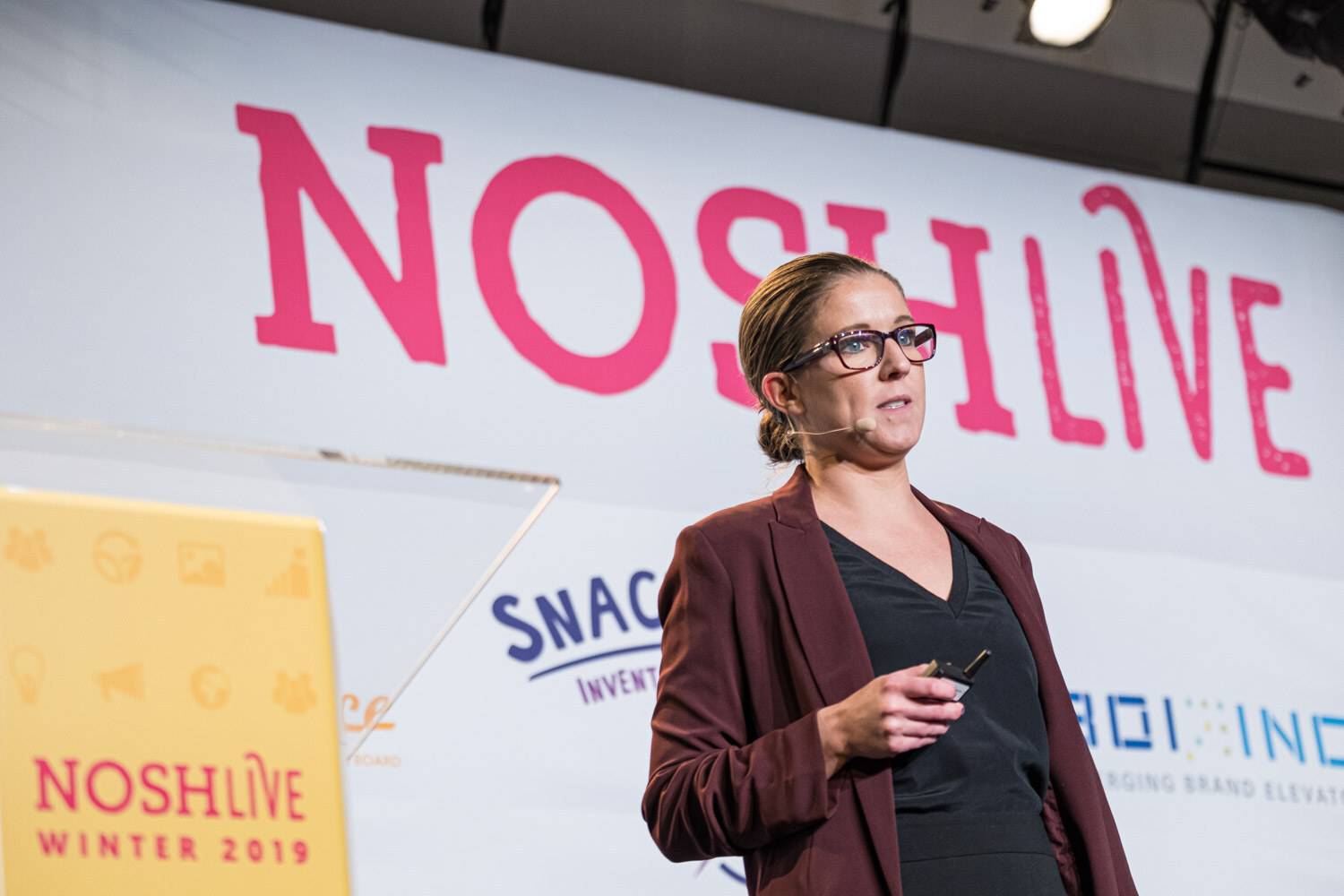A woman in a maroon blazer presenting beside a yellow Nosh Live lectern