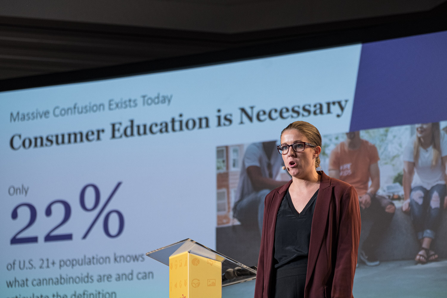 A woman speaking on stage in front of a slide reading Consumer Education is Necessary and a statistic of 22 percent