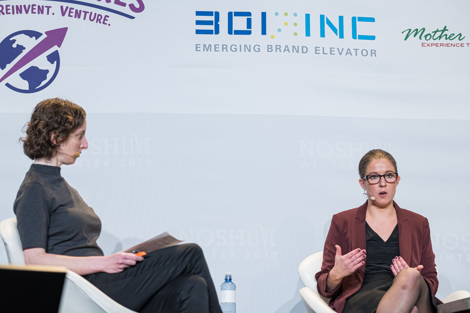 A woman in a maroon blazer gesturing while seated across from a moderator in a gray top