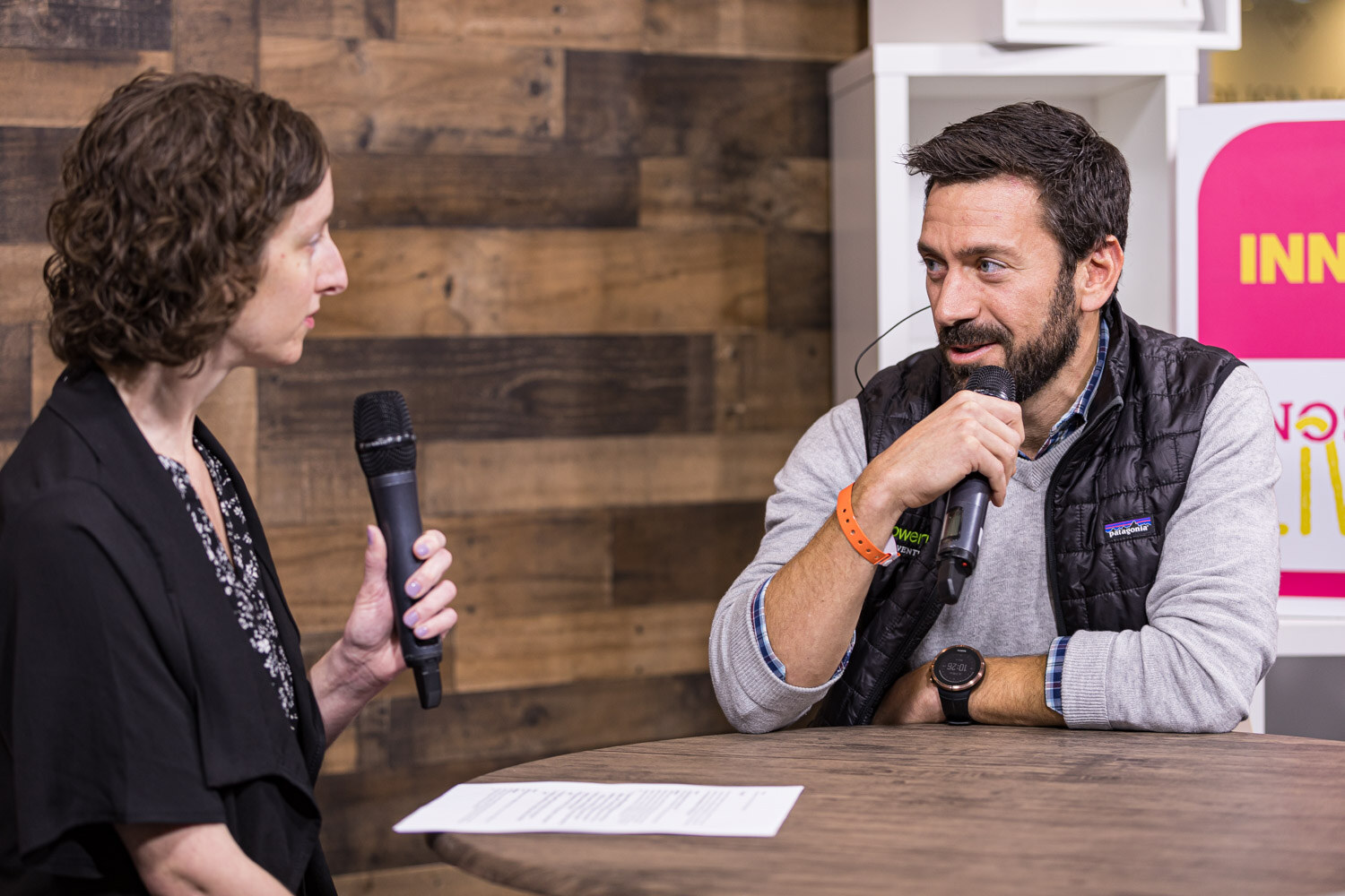 An interviewer holding a microphone talks with a man in a black vest at a round table in front of an Innovation sign
