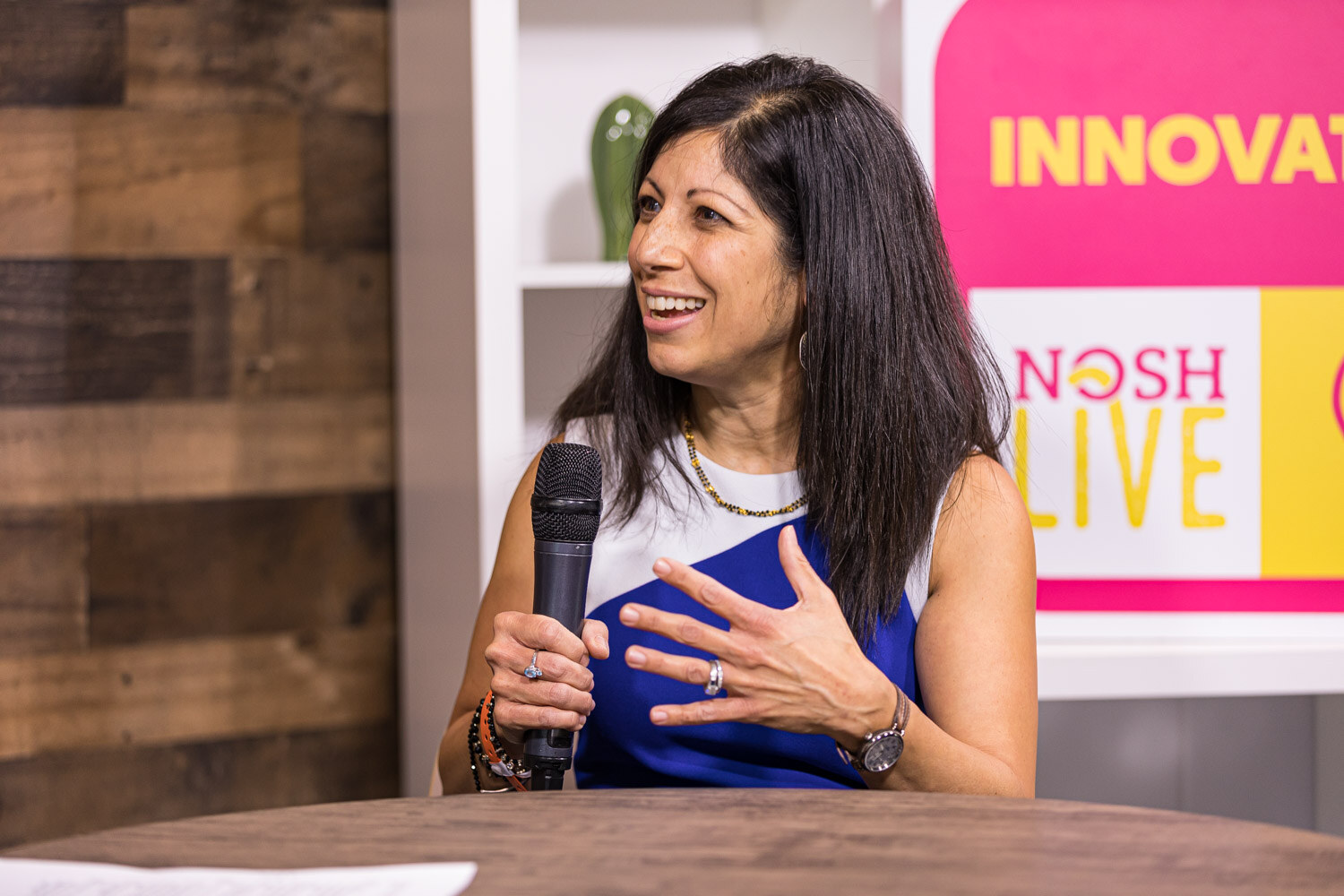 A smiling woman with long dark hair in a blue and white top holding a microphone at the Innovation set