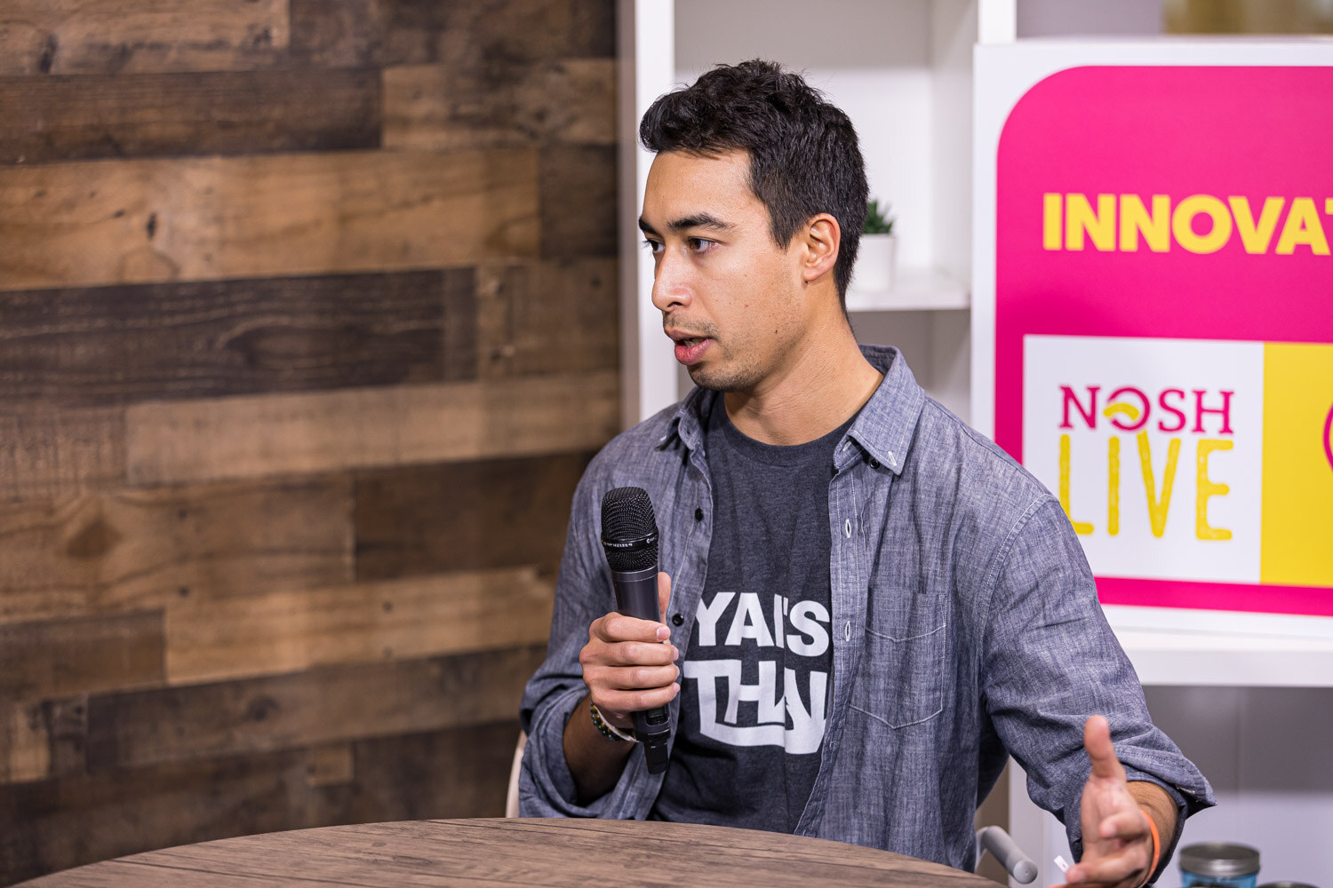 A man in a gray shirt over a graphic tee speaks into a microphone while gesturing