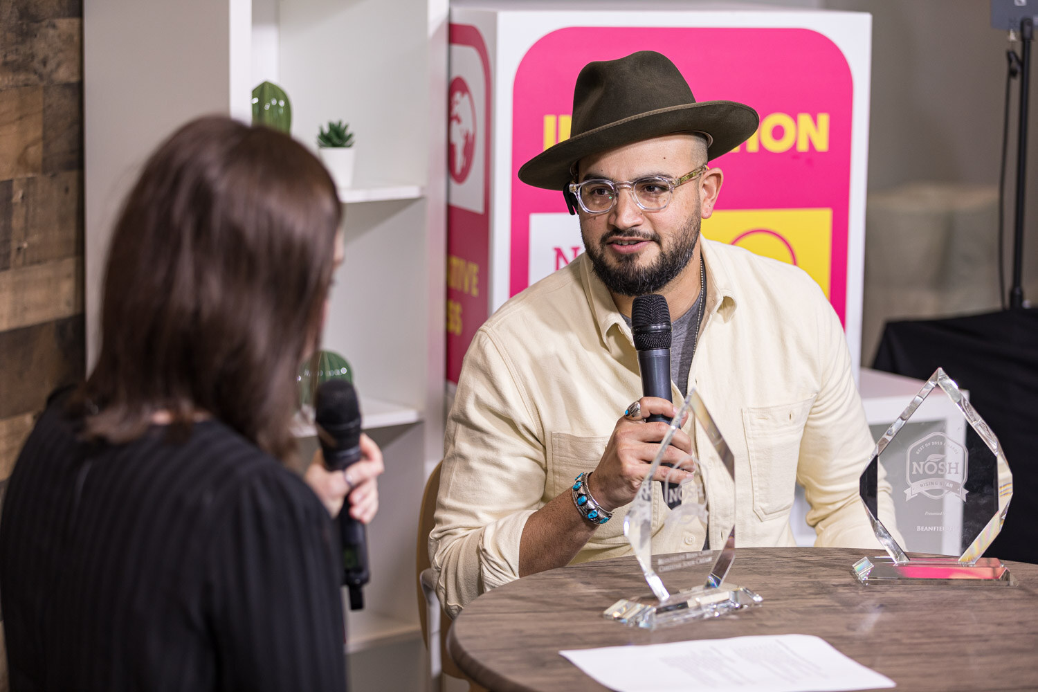 A man in a hat and glasses speaks into a microphone with two glass award trophies on the table in front of him