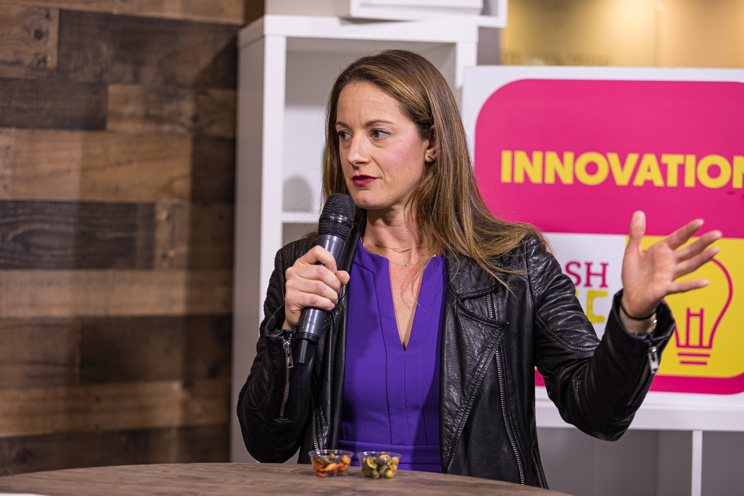 A woman in a black leather jacket gesturing as she talks, with small sample cups on the table