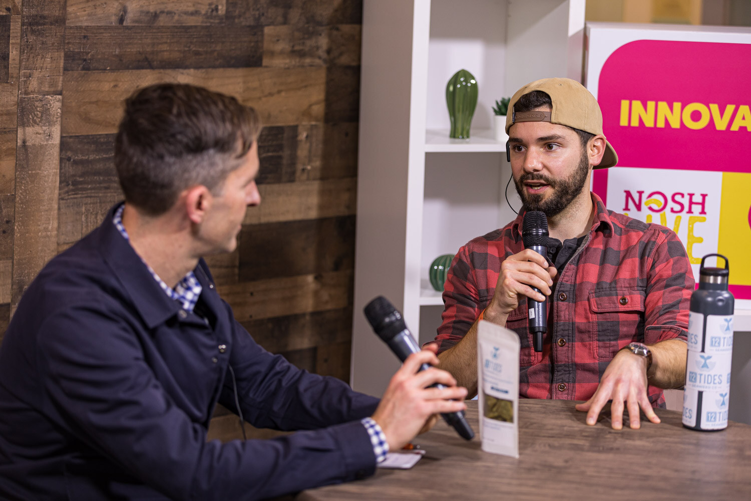 A man in a plaid shirt and cap speaks to an interviewer in a navy jacket across a round table