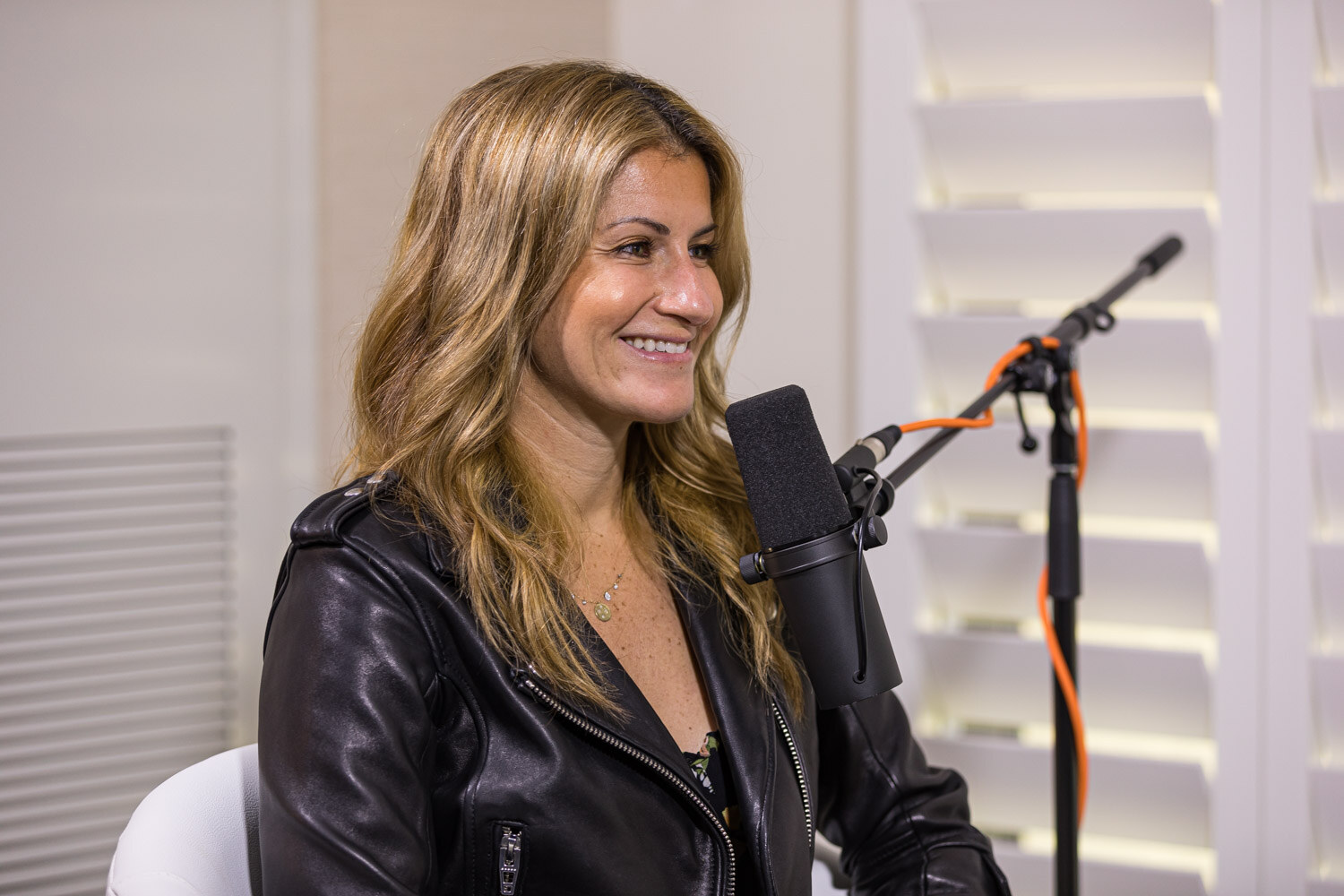A smiling woman in a black leather jacket seated beside a studio microphone in front of white shutters