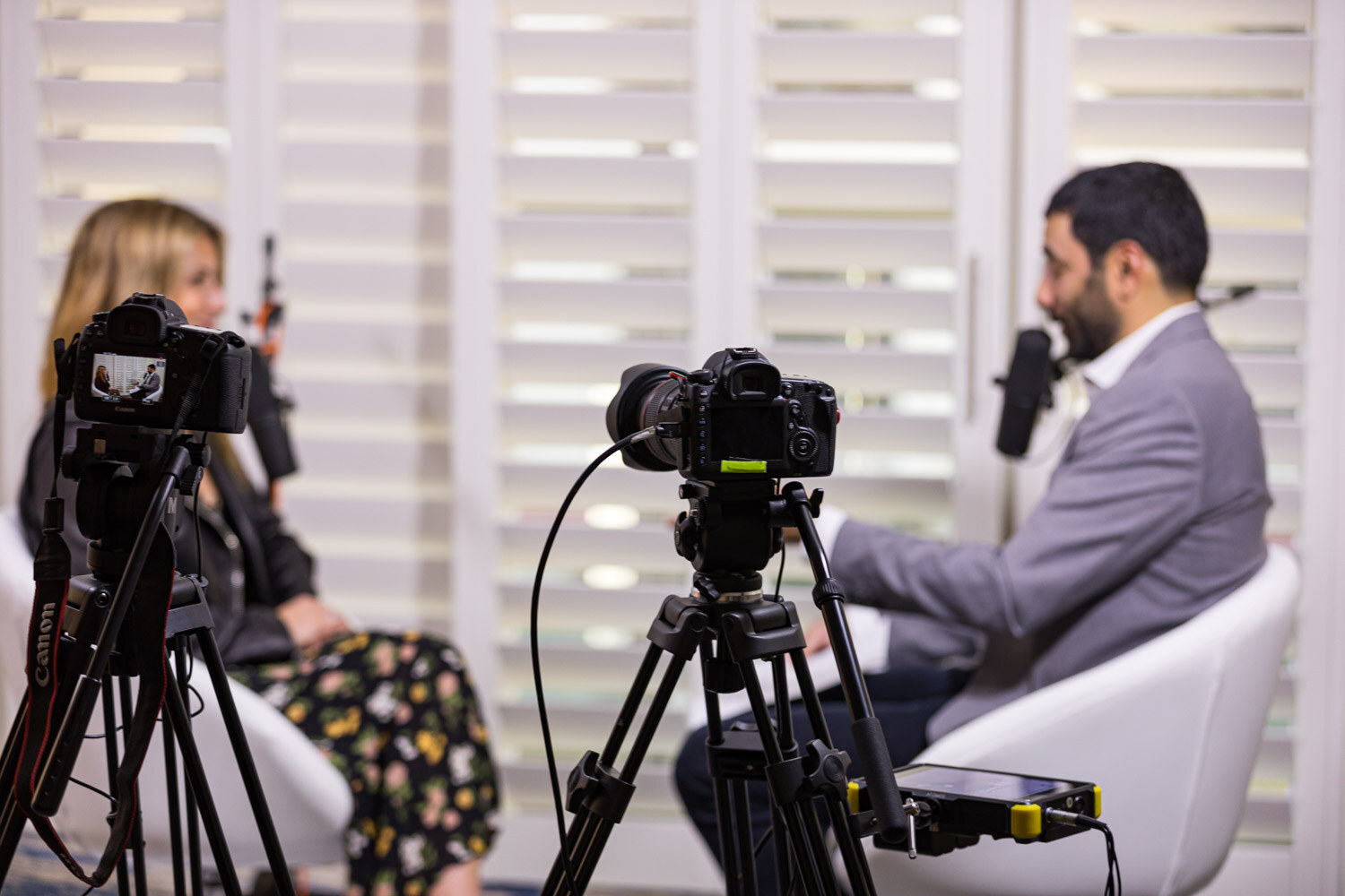 Two cameras on tripods film an interview between a seated woman and a man in a gray jacket