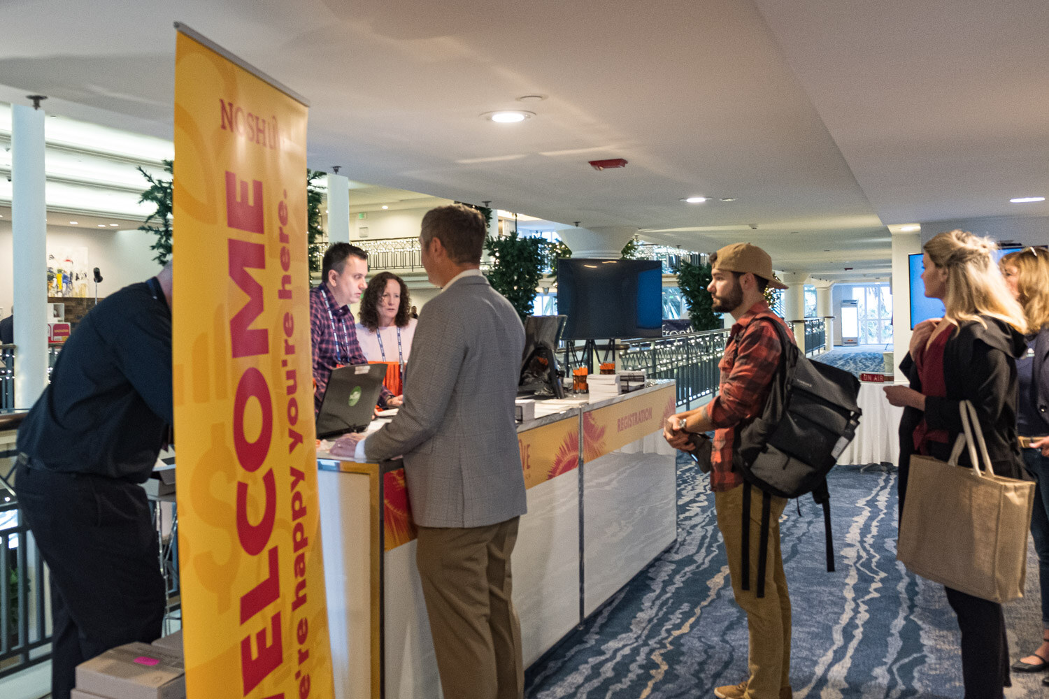 Attendees waiting at the registration counter beside a yellow Welcome, We're happy you're here banner
