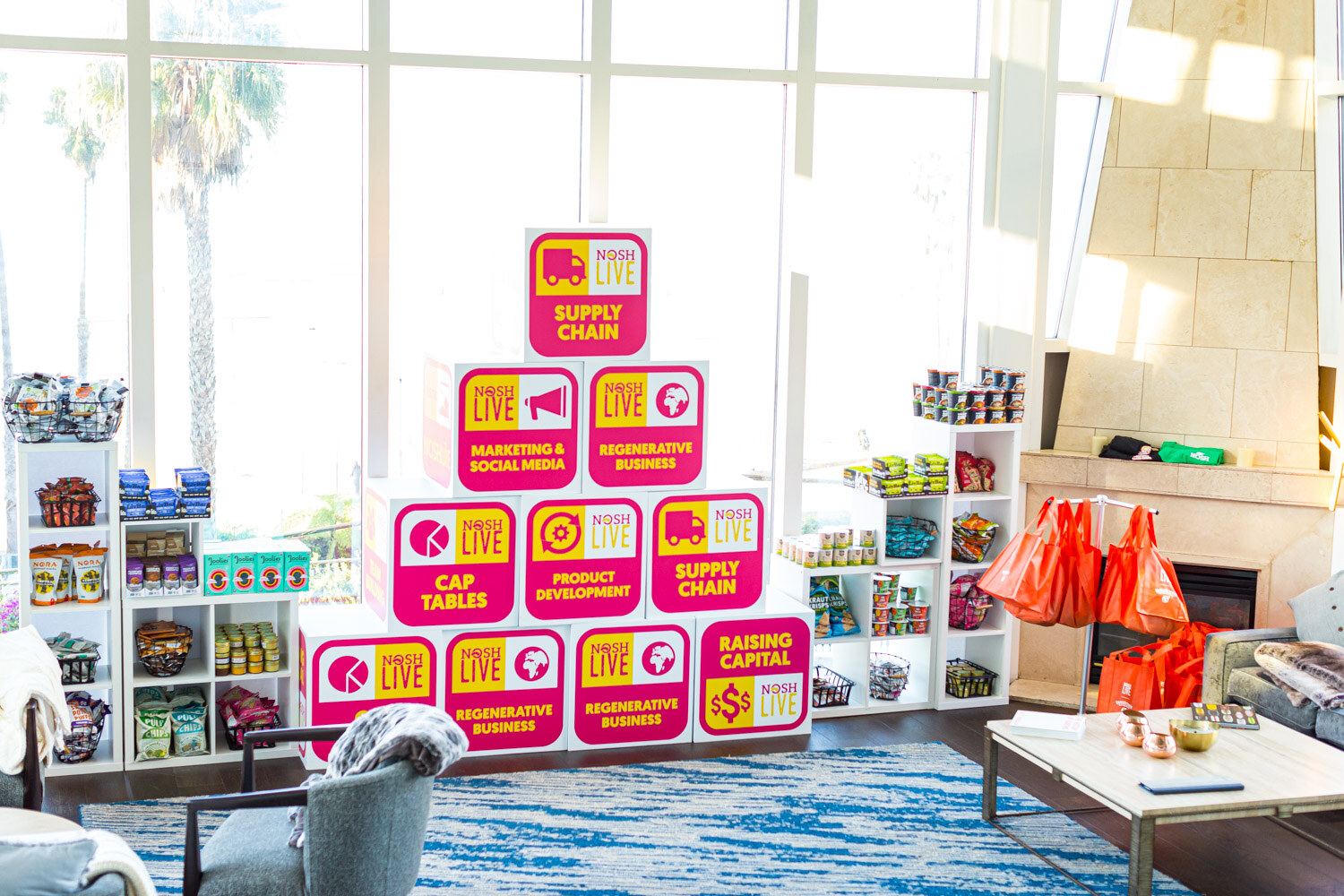 Stacked pink Nosh Live cubes labeled Supply Chain, Cap Tables and Raising Capital beside shelves of snack products in a bright room by a fireplace