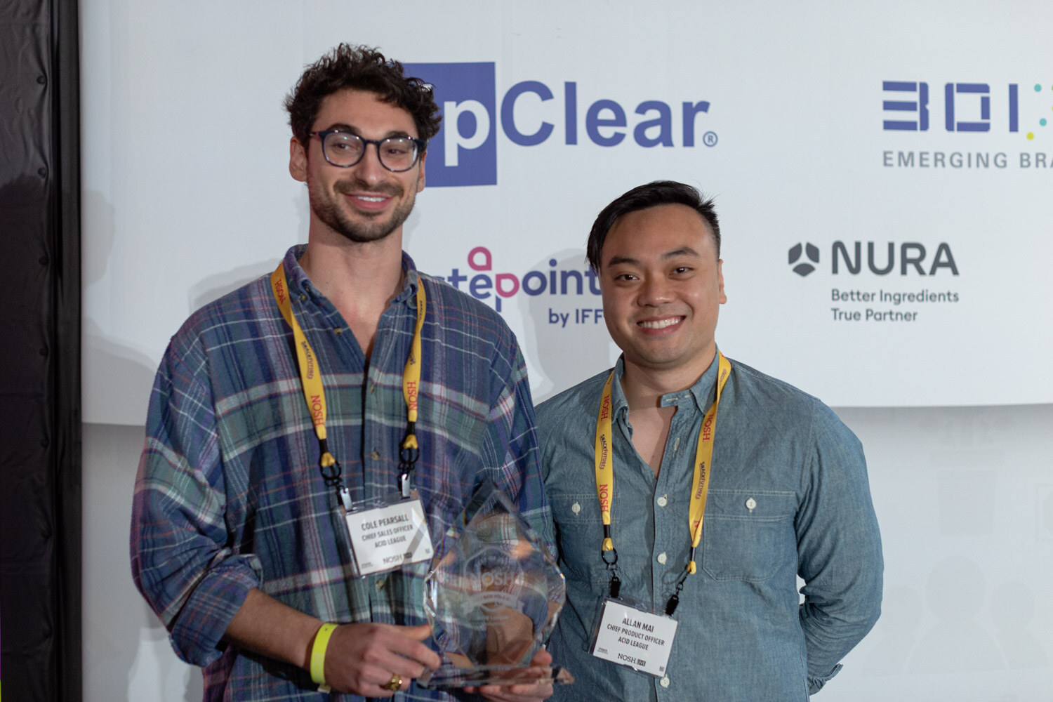 Man in a plaid shirt holding a glass Nosh award next to a man in a blue shirt, both wearing yellow Nosh lanyards, in front of a sponsor backdrop.