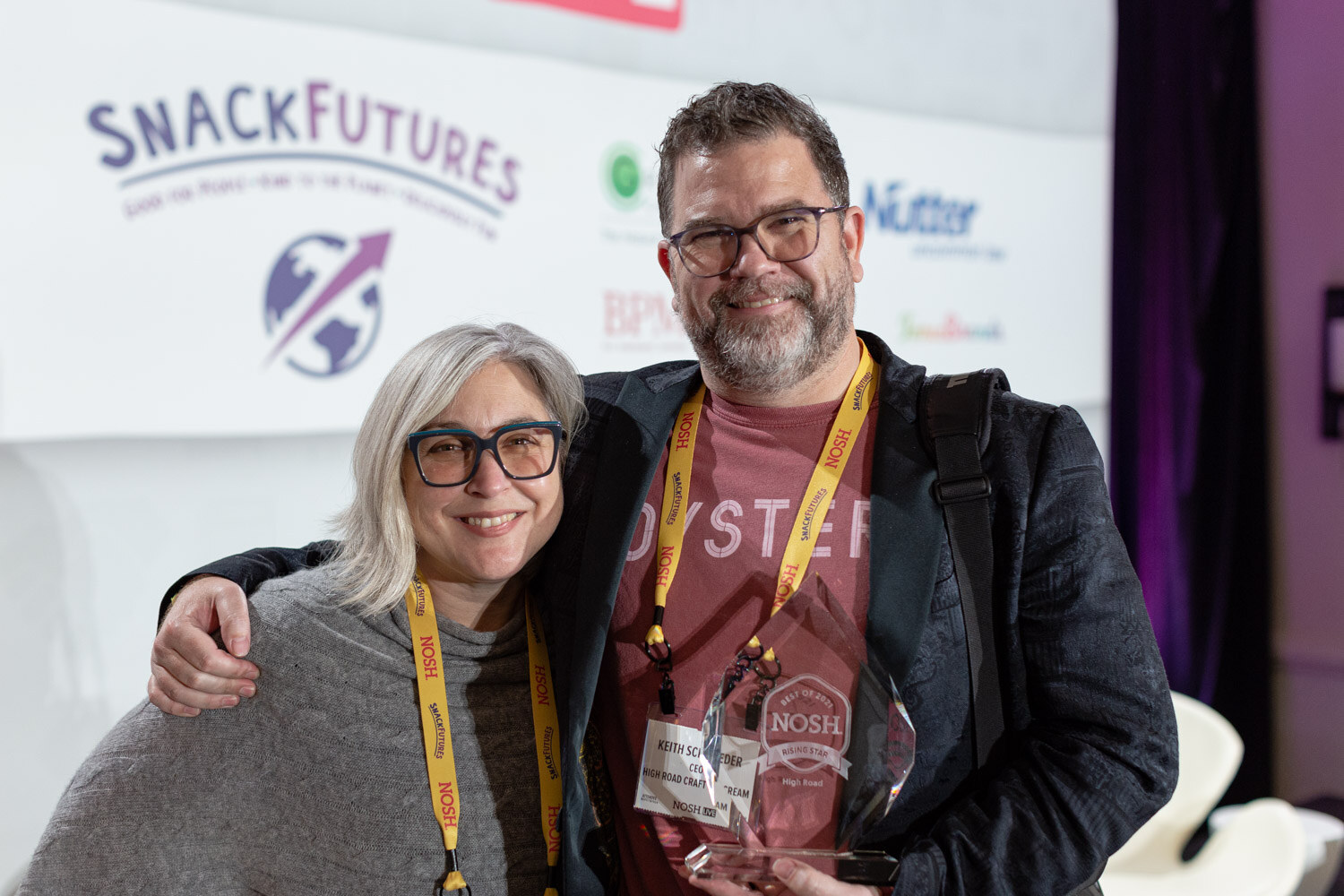 Bearded man in a dark jacket and red t-shirt with his arm around a woman with silver hair and glasses, holding a glass Nosh award in front of a SnackFutures backdrop.