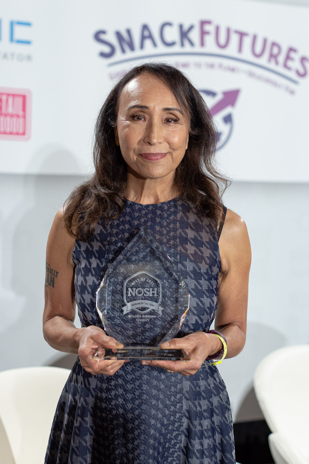Woman with long dark hair in a sleeveless navy houndstooth dress holding a glass Nosh award with both hands in front of a SnackFutures sponsor backdrop.