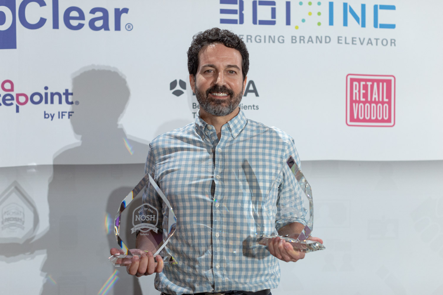 Bearded man in a blue gingham shirt holding two glass Nosh awards in front of a backdrop with UpClear, 301 Inc and Retail Voodoo logos.