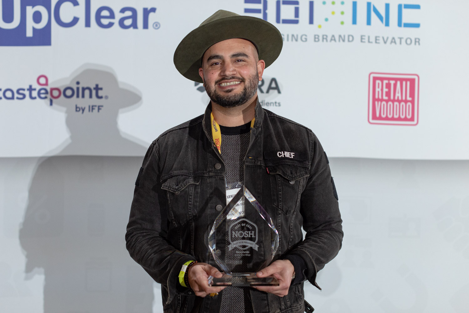 Smiling man with a beard and a green wide-brim hat in a black denim jacket holding a glass Nosh award in front of UpClear, Tastepoint and Retail Voodoo logos.