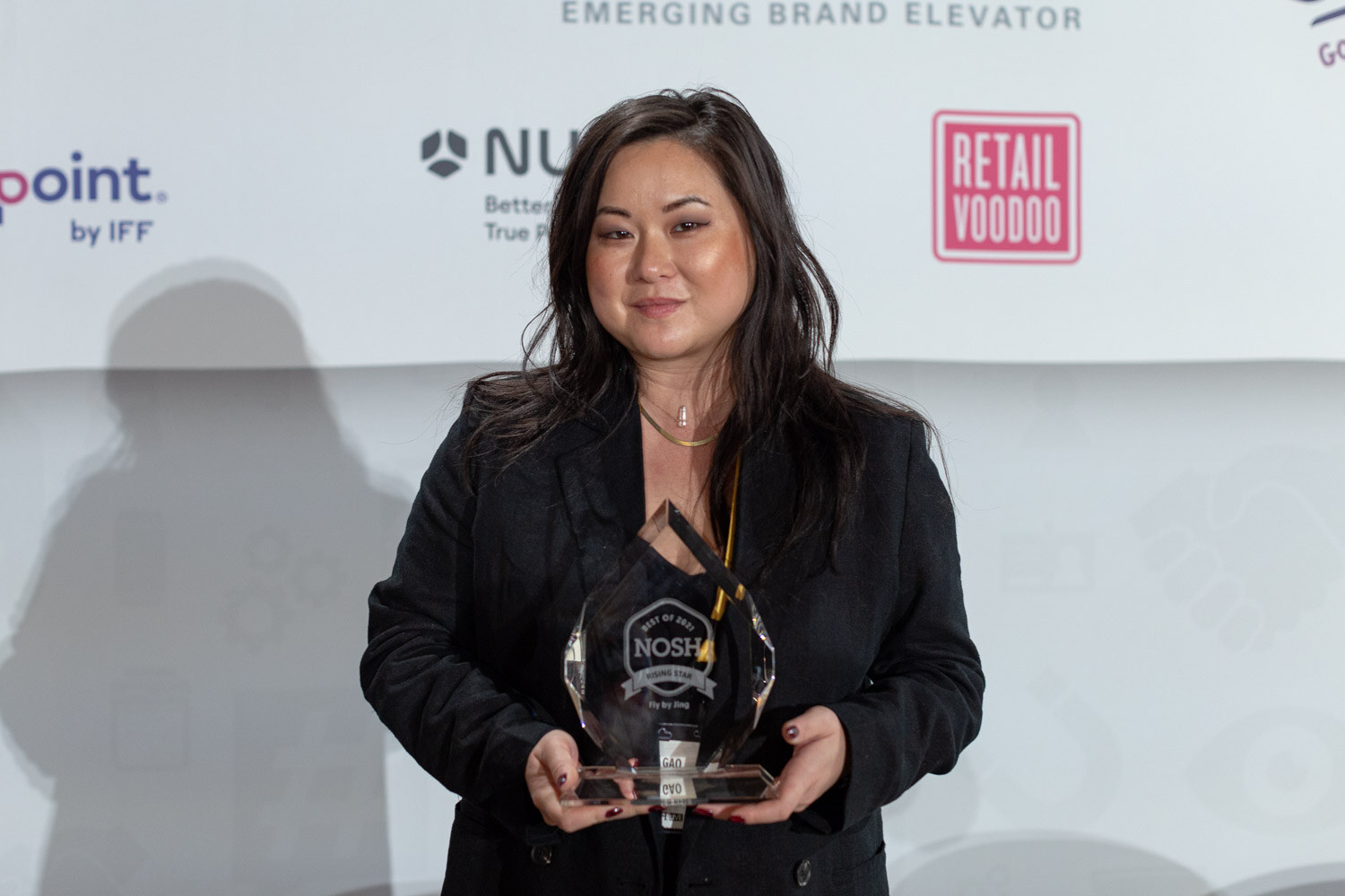 Woman with long dark hair in a black blazer holding a glass Nosh award engraved Rising Star in front of Retail Voodoo and NURA logos.