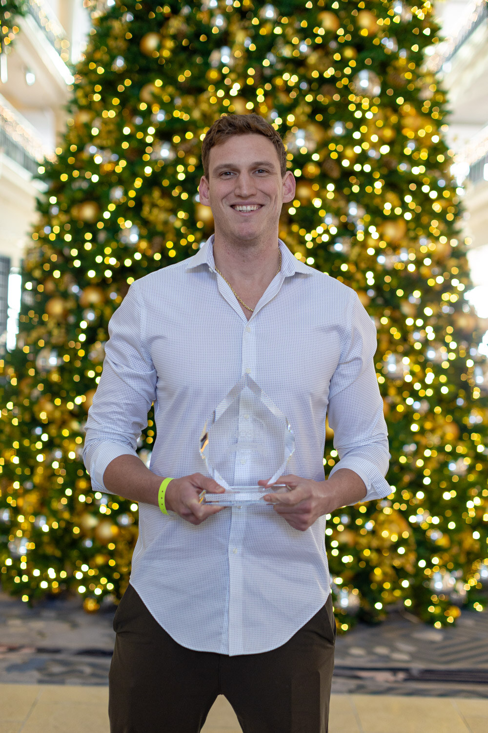 Smiling man in a white button-up shirt holding a clear glass award in front of a tall Christmas tree with warm yellow lights.