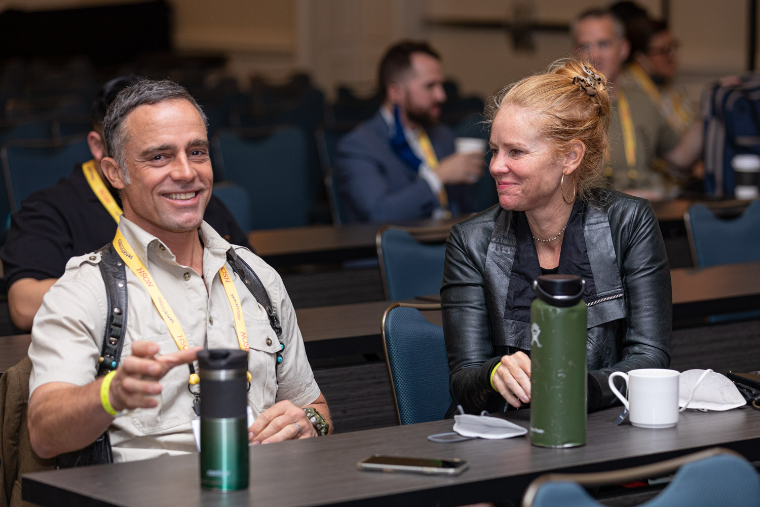 Man in a khaki shirt and a Nosh lanyard pointing toward the camera and a woman in a black leather jacket beside him, at a long table with water bottles and masks.