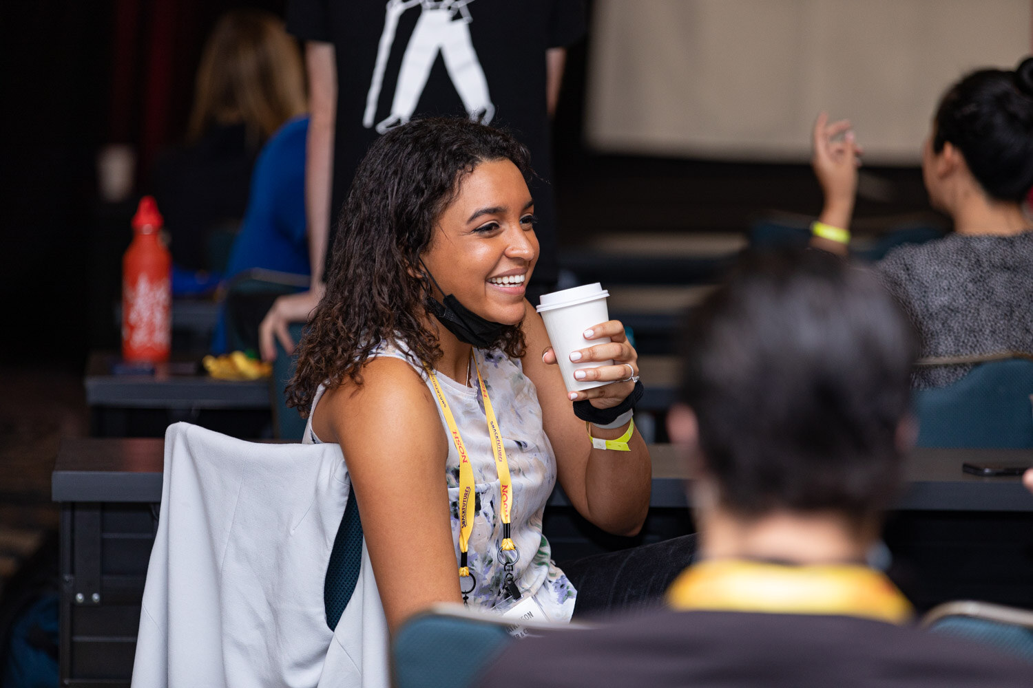 Smiling woman with curly dark hair in a sleeveless top and yellow Nosh lanyard holding a white coffee cup, with a mask pulled below her chin, seated in the ballroom.