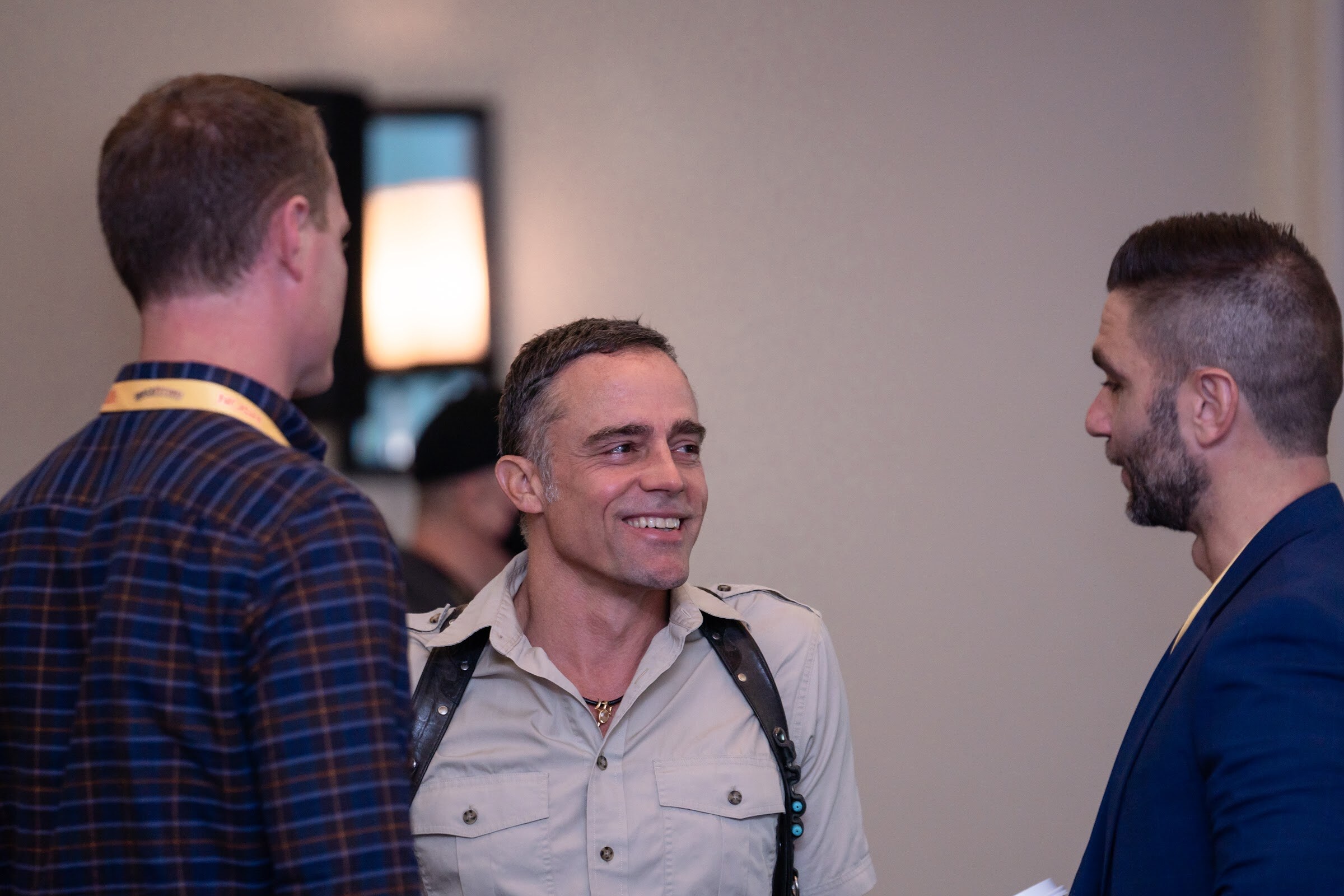 Man in a khaki shirt with shoulder straps smiling between a man in a plaid shirt with a yellow lanyard and a man in a navy blazer.