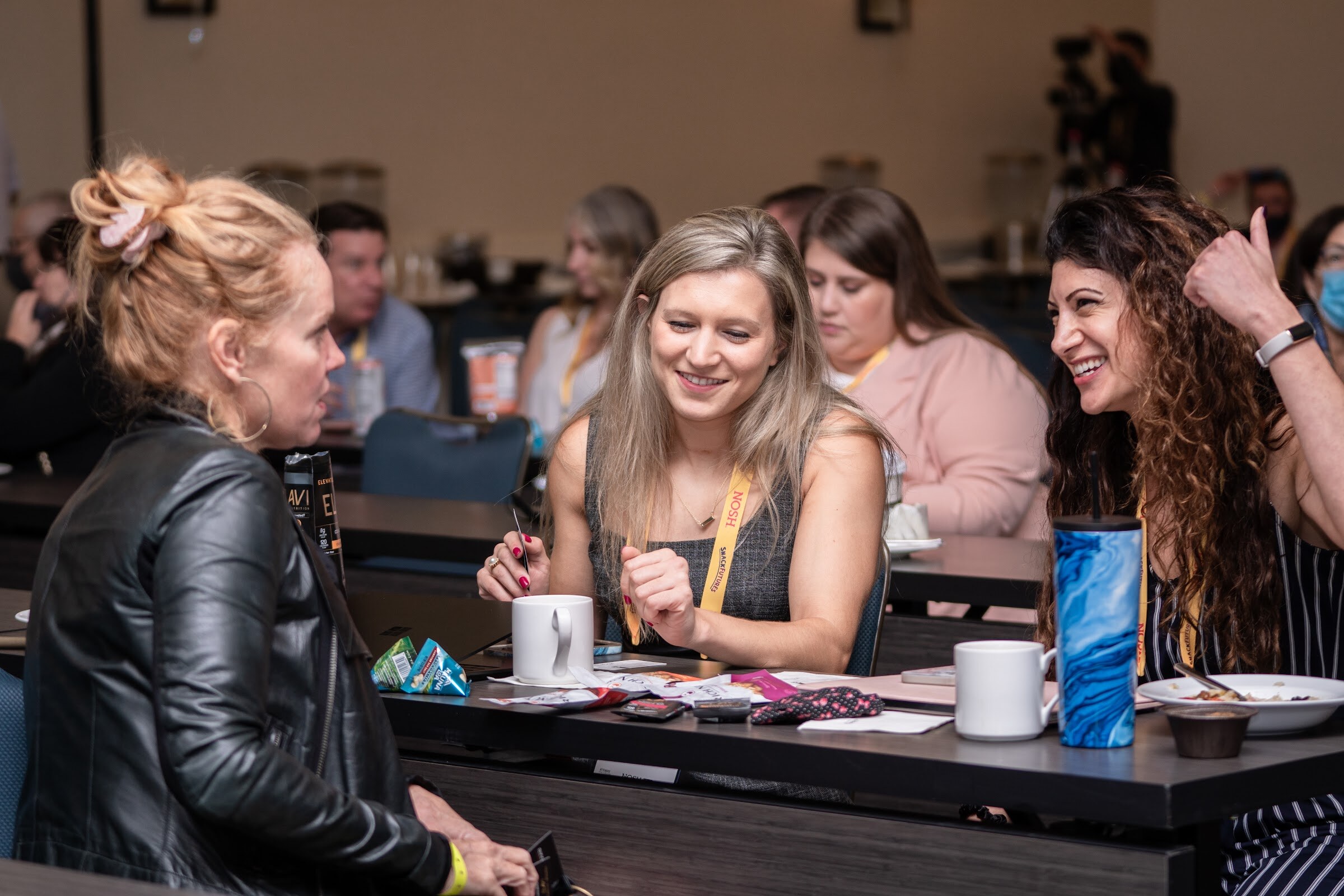 Woman with blonde hair in a grey top and woman with long curly dark hair, both in yellow lanyards and smiling, with a woman in a black leather jacket at the left, at a long table with mugs and snacks.