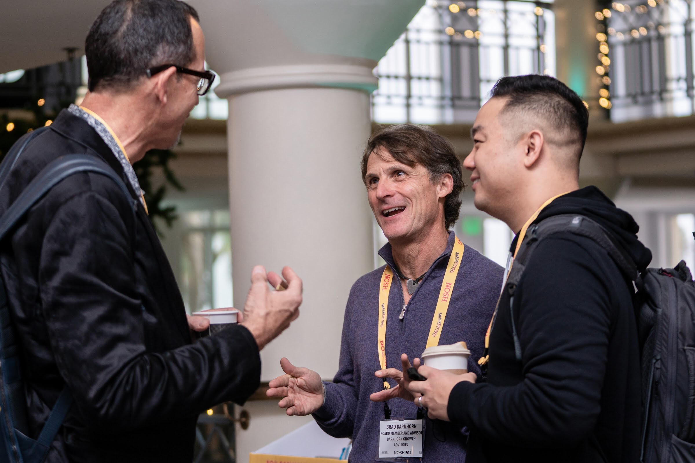 Three men in yellow Nosh lanyards standing near a white column in a bright atrium, one in a black jacket turned away and two facing him and smiling.