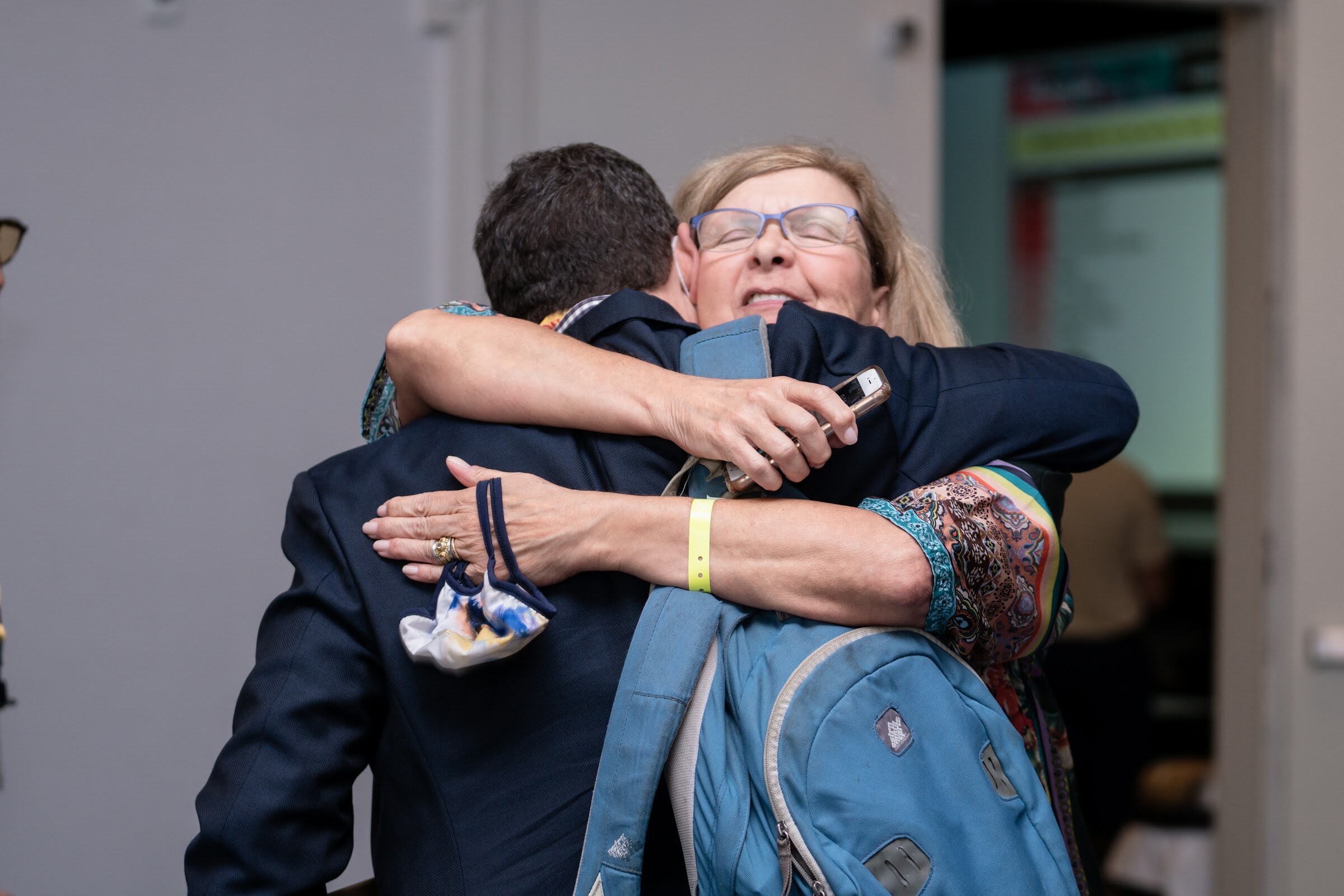 A man in a navy blazer and a woman with glasses embracing, with a blue backpack and a yellow wristband visible.