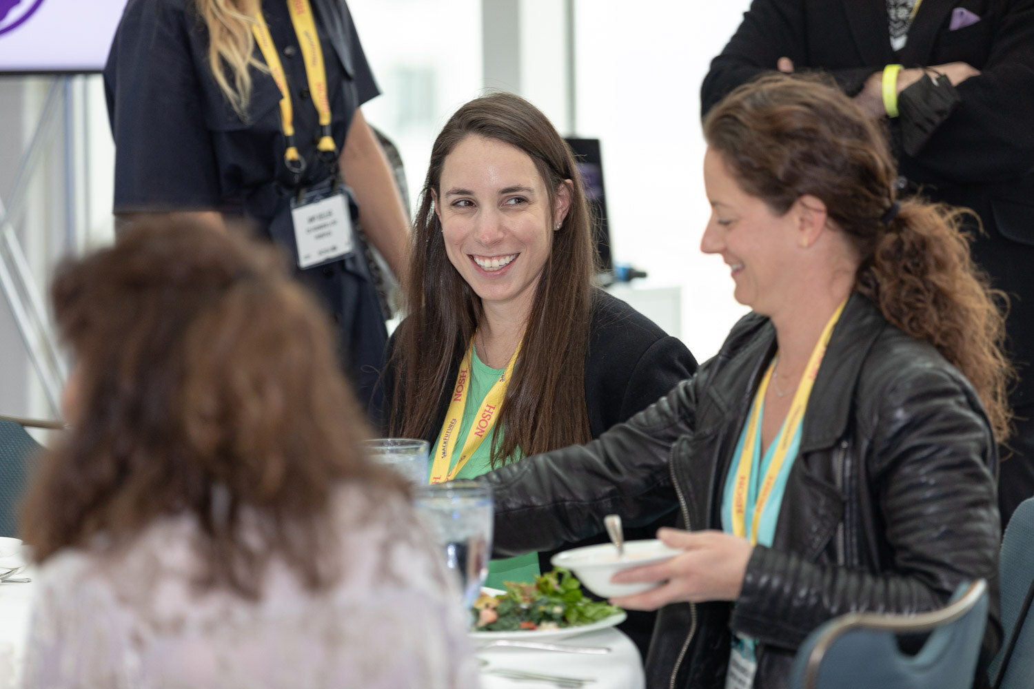 Woman with long brown hair in a green top and woman in a black leather jacket, both with yellow Nosh lanyards, laughing at a table with plates of salad.