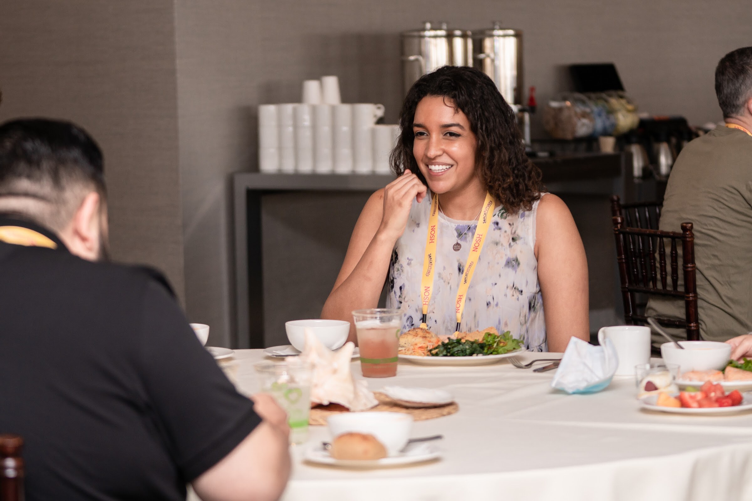 Smiling woman with curly dark hair in a sleeveless floral top and a yellow Nosh lanyard at a round table set with plates of food and glasses.