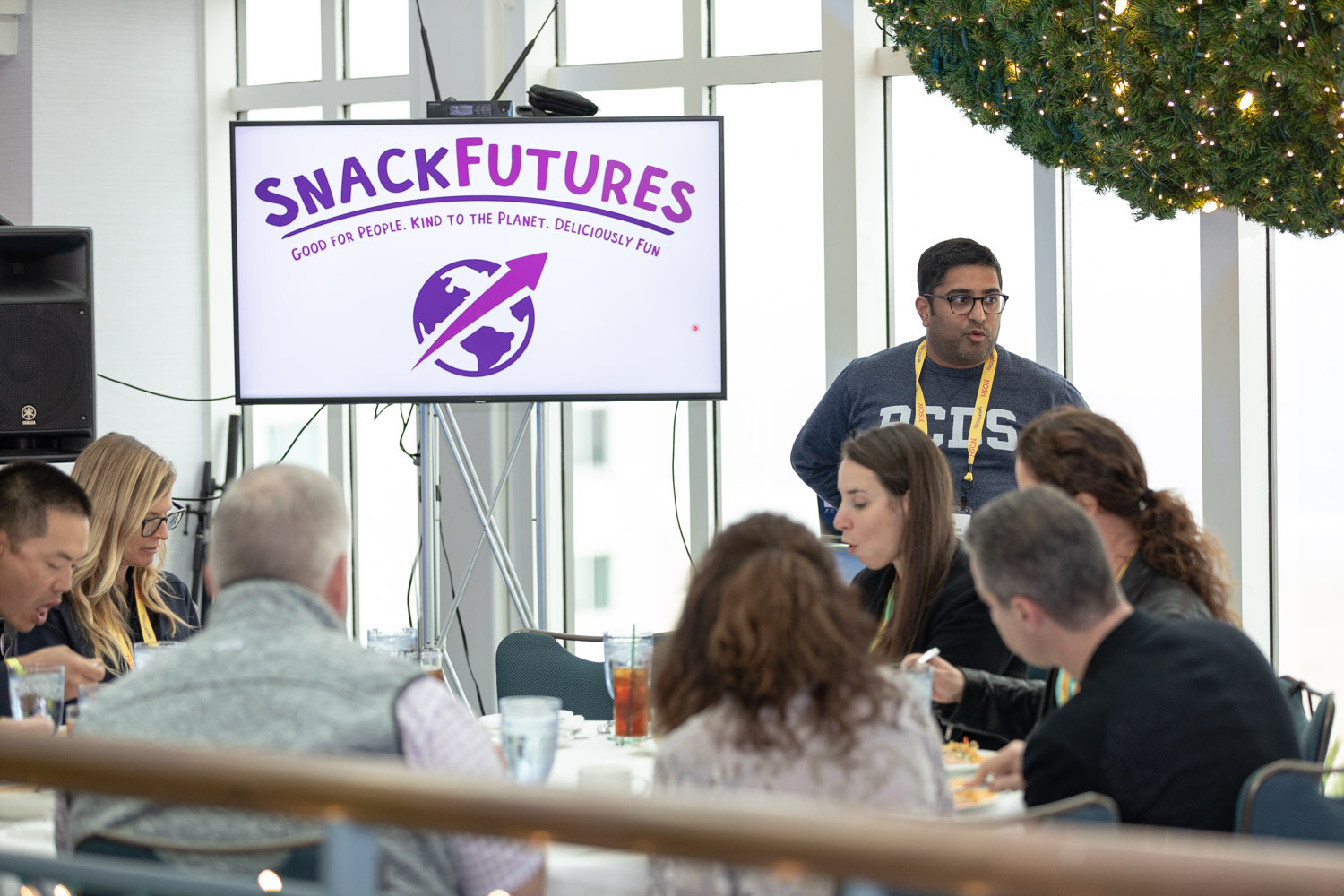 Room with a screen reading SnackFutures Good for People, Kind to the Planet, Deliciously Fun, a man in a navy sweatshirt standing behind seated attendees and a holiday wreath at the top right.