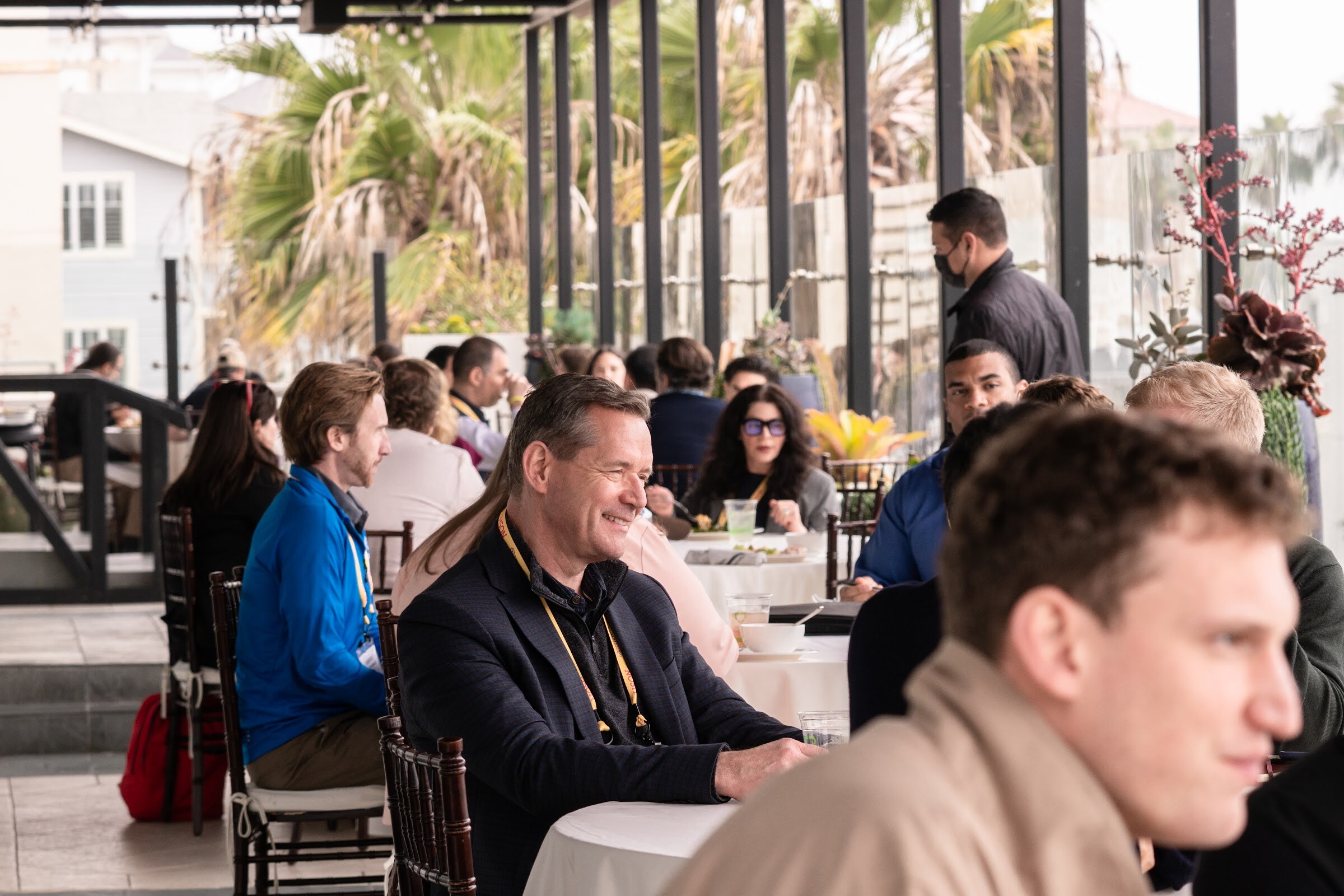 Crowded outdoor terrace with attendees in yellow Nosh lanyards seated at tables, a smiling man in a dark jacket in the foreground and palm trees in the background.