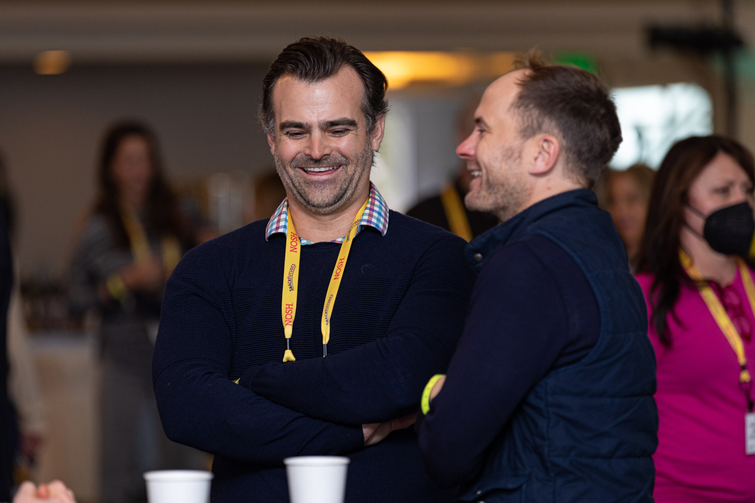 Man in a navy sweater with arms crossed and a Nosh lanyard smiling beside a man in a navy vest laughing, with other attendees in the background.