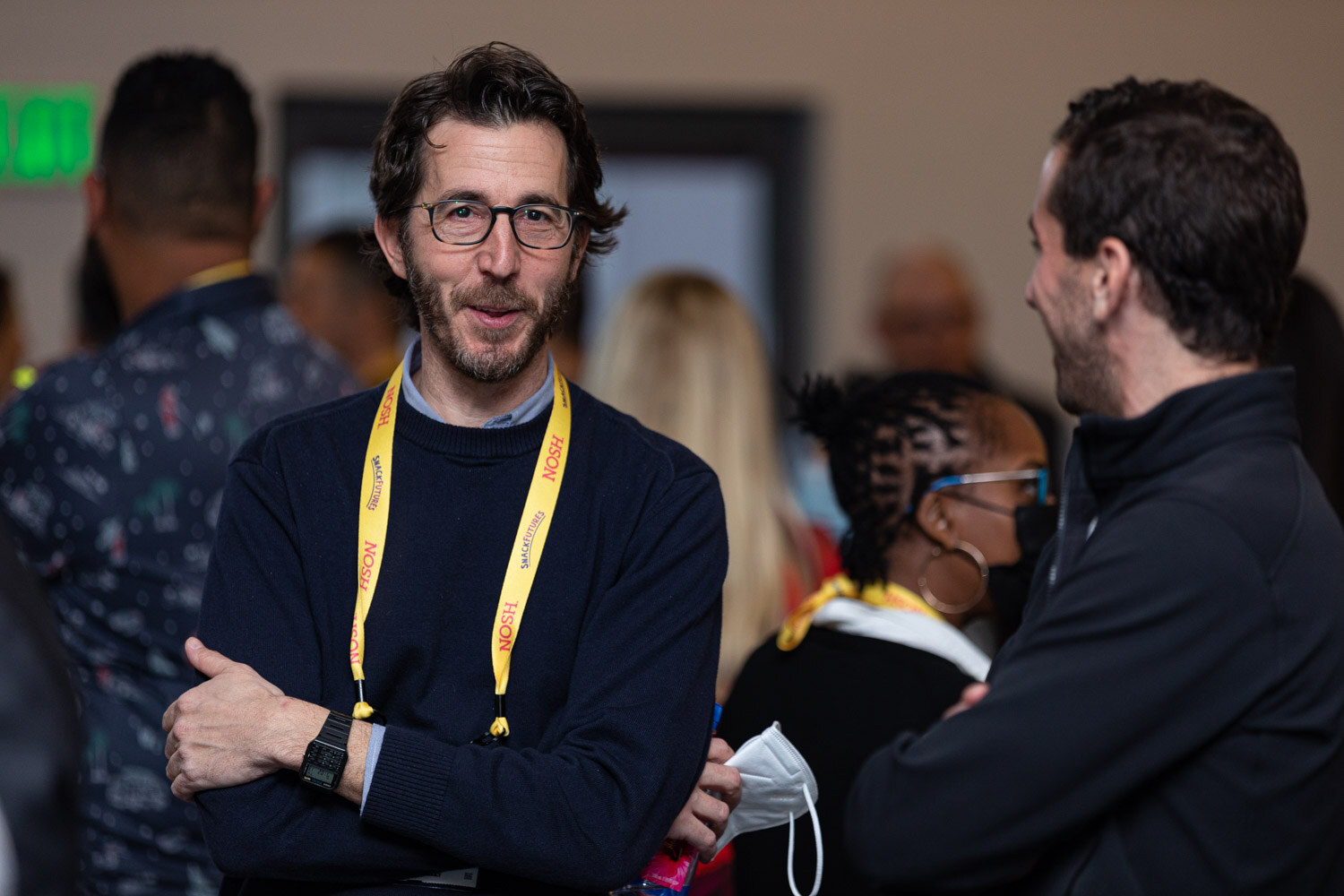 Man with dark curly hair, glasses and a beard in a navy sweater and a yellow Nosh lanyard with arms crossed, with attendees talking behind him.