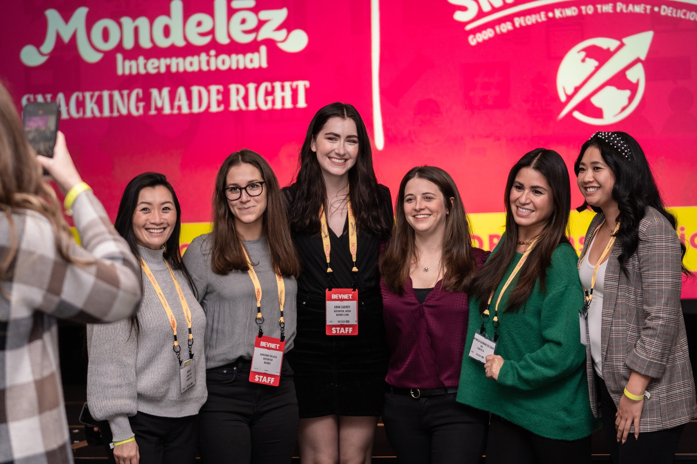 Six smiling women in a row in front of a pink screen reading Mondelez International Snacking Made Right, several wearing red BevNET staff badges and yellow Nosh lanyards, while another attendee photographs them on a phone.