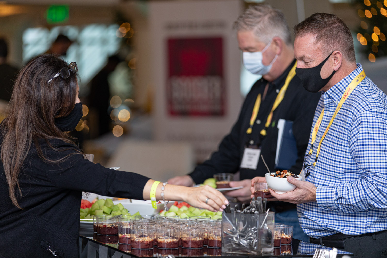 Woman reaching for a parfait cup, a man in a checked shirt holding a bowl and a man in a black shirt, all in masks and yellow Nosh lanyards, at a buffet table with fruit and parfaits.