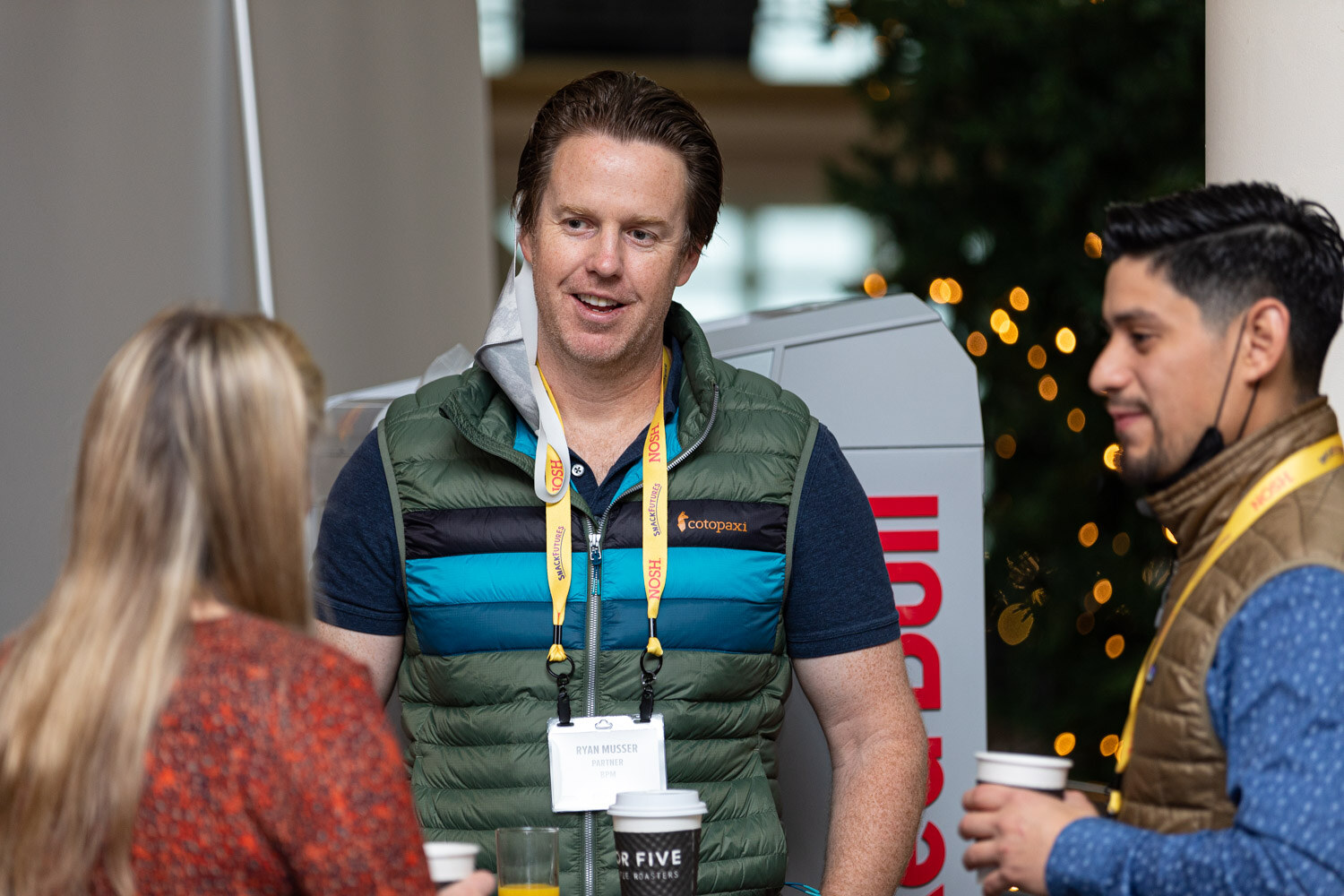 Man in a green puffer vest and Nosh lanyard smiling at a woman with her back to the camera, with another man in a brown vest and a lit tree in the background.