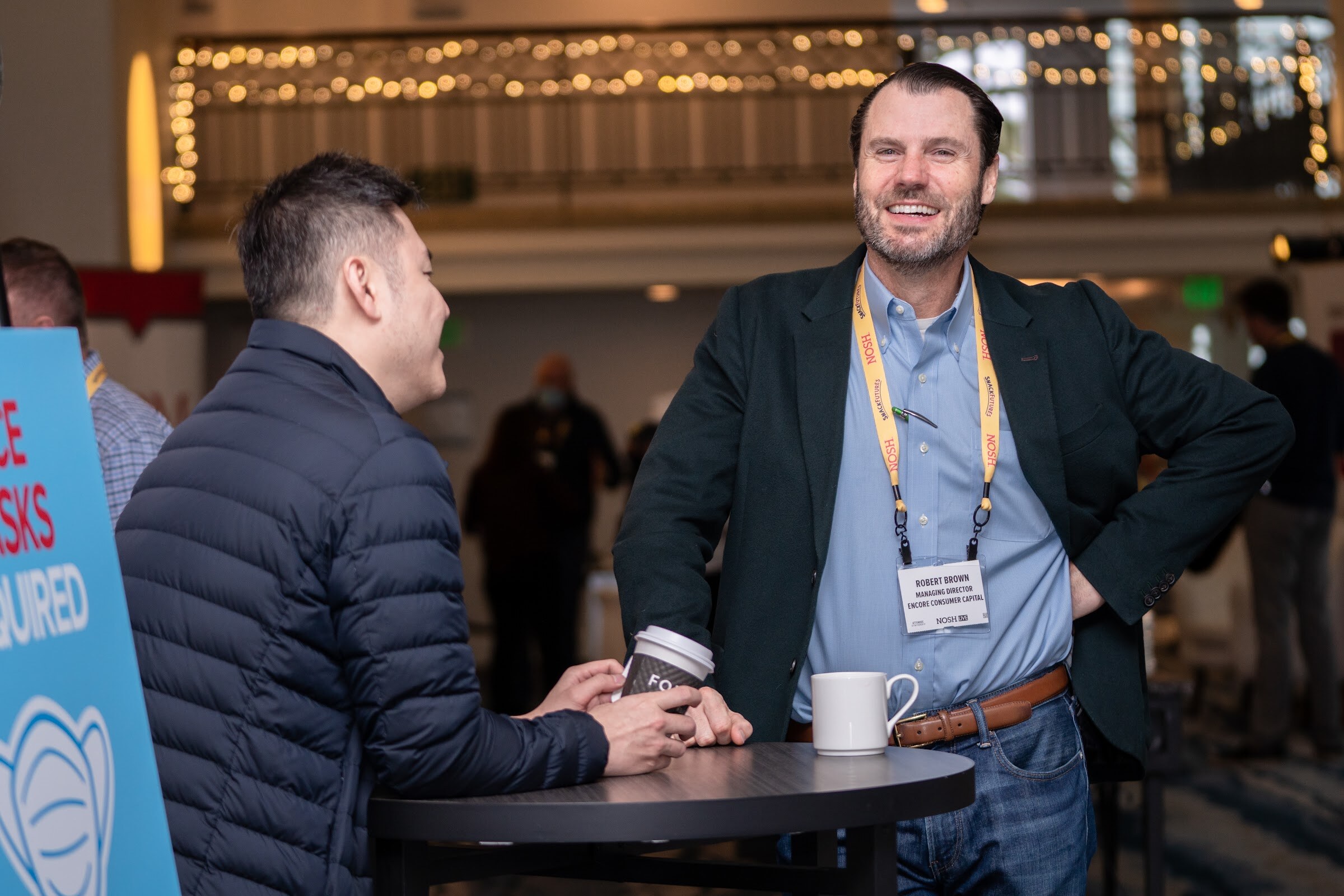 Man with a beard in a dark blazer and light blue shirt with a yellow Nosh lanyard leaning on a high-top table beside a man in a navy puffer jacket, with string lights on the balcony behind them.